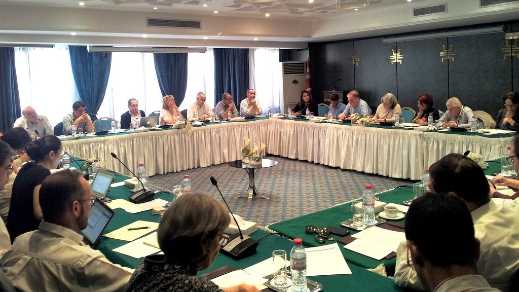 L'image montre une grande salle de réunion où se déroule une conférence ou un séminaire. Plusieurs personnes sont assises autour d'une table ovale, certaines utilisant des ordinateurs portables. Les participants semblent engagés dans des discussions, et il y a des documents et des bouteilles d'eau sur chaque table. La pièce est bien éclairée par la lumière naturelle qui entre par de grandes fenêtres, et des rideaux bleus ajoutent une touche décorative. Le décor est professionnel, avec des éléments modernes et une ambiance de collaboration.