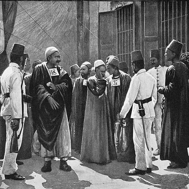 The image depicts a historical scene featuring a group of men, likely from a Middle Eastern or North African context. They are dressed in traditional attire, which includes long robes and distinctive hats. Some men are holding numbered placards, suggesting they may be part of a formal event or a historical gathering. The setting appears to be outdoors, possibly in an urban environment, with walls and structures in the background. The expressions and postures of the individuals suggest a moment of interaction or discussion among the group.