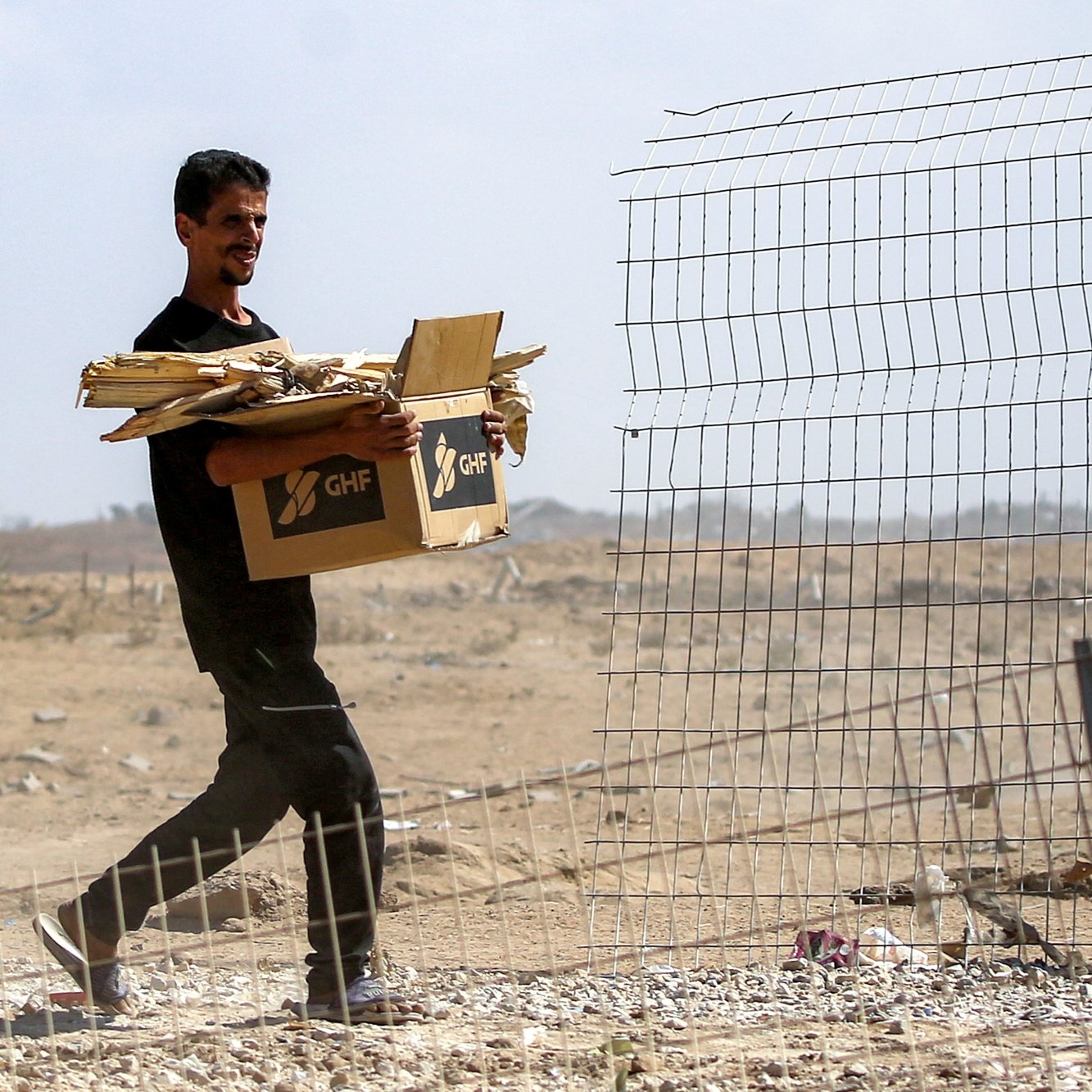 Homme portant une boîte et du bois près d'une clôture dans un paysage désertique.