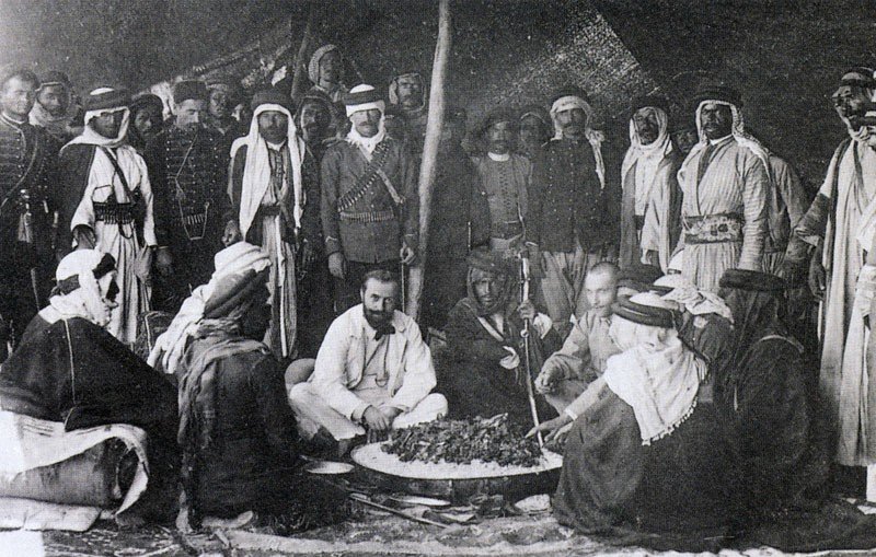 این تصویر تاریخی گروهی از مردان را در یک محیط سنتی نشان می‌دهد. آنها در یک چادر یا فضای باز گرد هم آمده‌اند و به نظر می‌رسد در حال تهیه یا صرف غذا هستند. چند نفر از آن‌ها لباس‌های سنتی عربی به تن دارند، از جمله عبا و headscarf (کشیه). در مرکز تصویر، چند نفر در حال نشستن و گفتگو هستند و سایرین ایستاده یا نشسته در اطراف آن‌ها قرار دارند. فضای تصویر گرم و صمیمی به نظر می‌رسد و نمایانگر فرهنگی غنی و سنت‌های اجتماعی است.