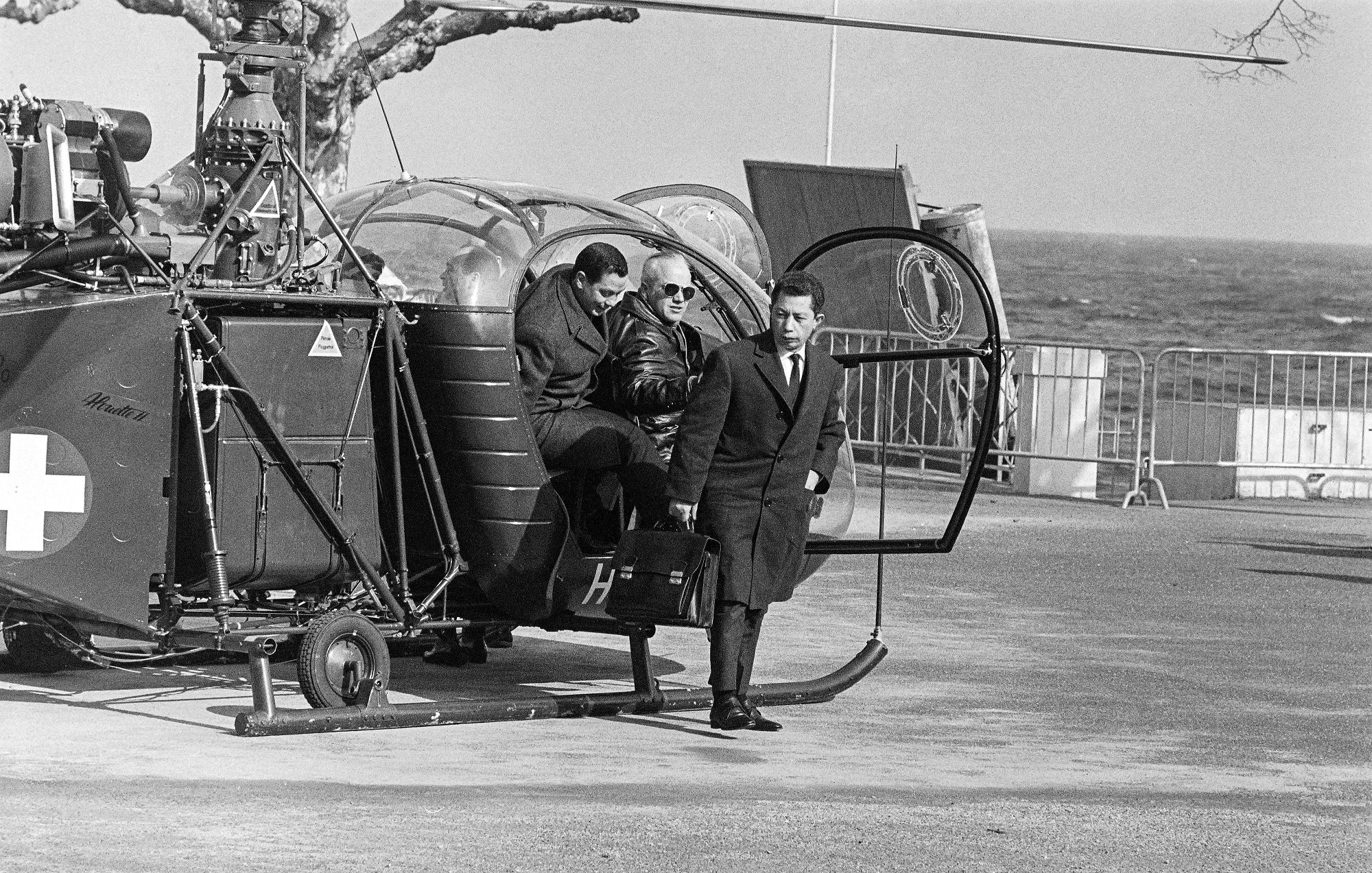 L'image montre une scène en noir et blanc avec un hélicoptère garé près de la mer. Deux personnes descendent de l'hélicoptère, tandis qu'une troisième personne les attend. On peut les voir porter des vêtements formels, ce qui suggère une certaine occasion. L'environnement est dégagé, avec une barrière visible en arrière-plan et des arbres dénudés. La mer se trouve en arrière-plan, ajoutant une touche de paysage côtier à la scène.