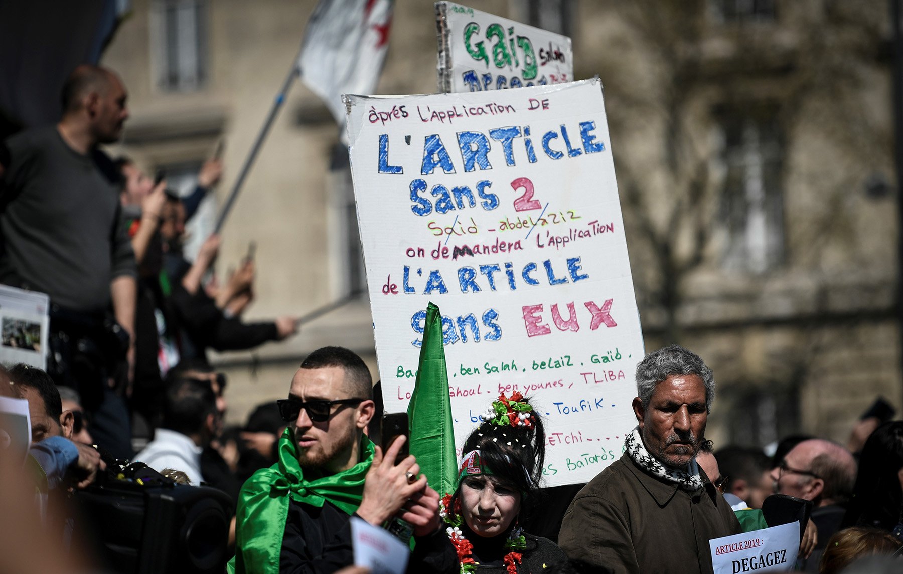 L'image montre une manifestation avec des participants tenant des pancartes. L'une des pancartes est bien visible et contient du texte en français, notamment une référence à l'article 2 et des demandes concernant cet article. Les manifestants portent des vêtements aux couleurs de l'Algérie, ce qui suggère un lien avec des préoccupations politiques ou sociales. L'atmosphère semble sereine mais déterminée, avec des gens rassemblés pour exprimer leurs revendications.