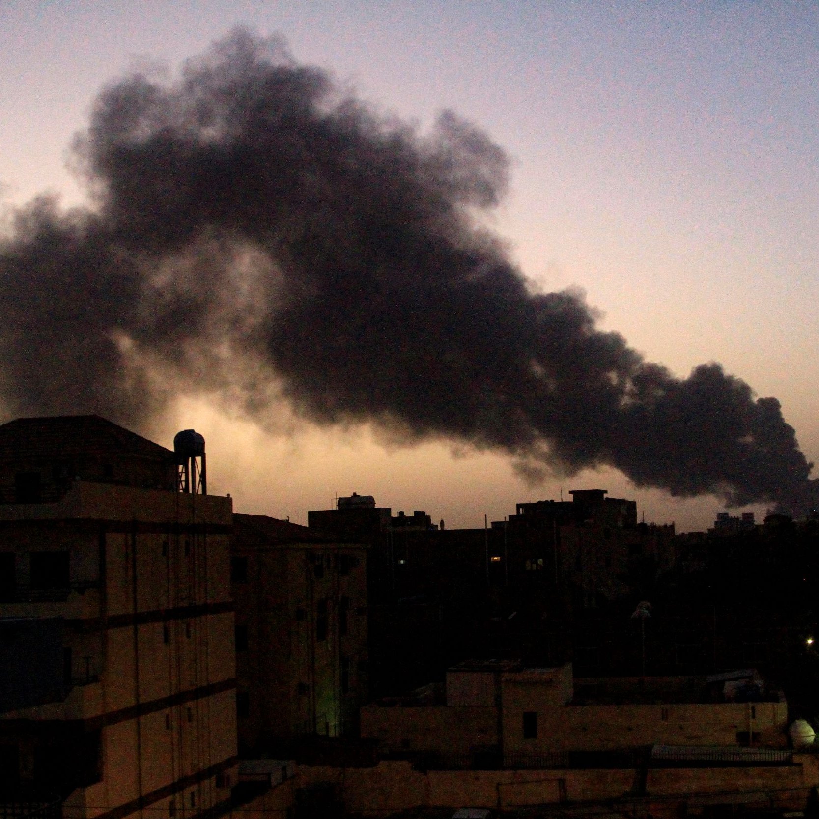 تظهر الصورة مشهداً لسماء غامقة تتخللها غيوم من الدخان الأسود تتصاعد في الأفق. تبدو المباني في المقدمة بلون داكن، مما يعكس ظلام الوقت الذي قد يكون عند الغسق. بينما تضفي أشجار النخيل لمسة خضراء في الجانب. الدخان يبدو كثيفاً وواضحاً، مما يشير إلى وجود حريق أو حدث صناعي في المنطقة.