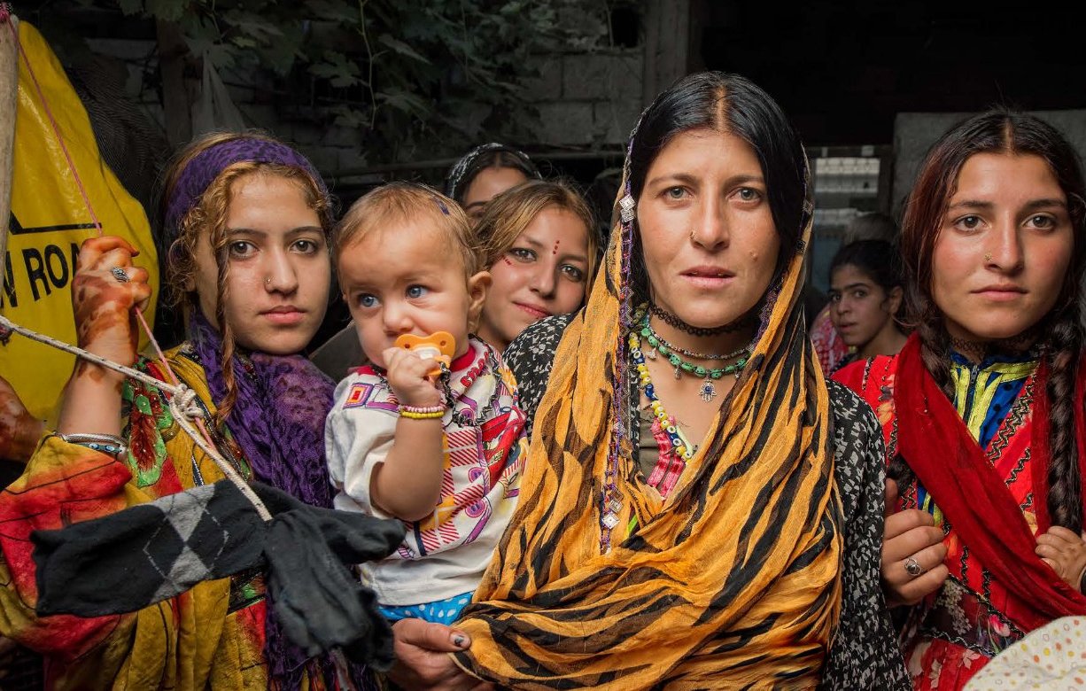 L'image montre un groupe de femmes et d'enfants, probablement dans un environnement rural. Les femmes portent des vêtements colorés et traditionnels, avec des foulards sur la tête. L'une d'elles tient un bébé dans ses bras. Les expressions sur leurs visages sont variées, allant de la curiosité à la détermination. On peut aussi voir des éléments culturels dans leur tenue, renforçant l'idée d'une riche tradition. L'arrière-plan semble un peu flou, suggérant un cadre rural ou communautaire.