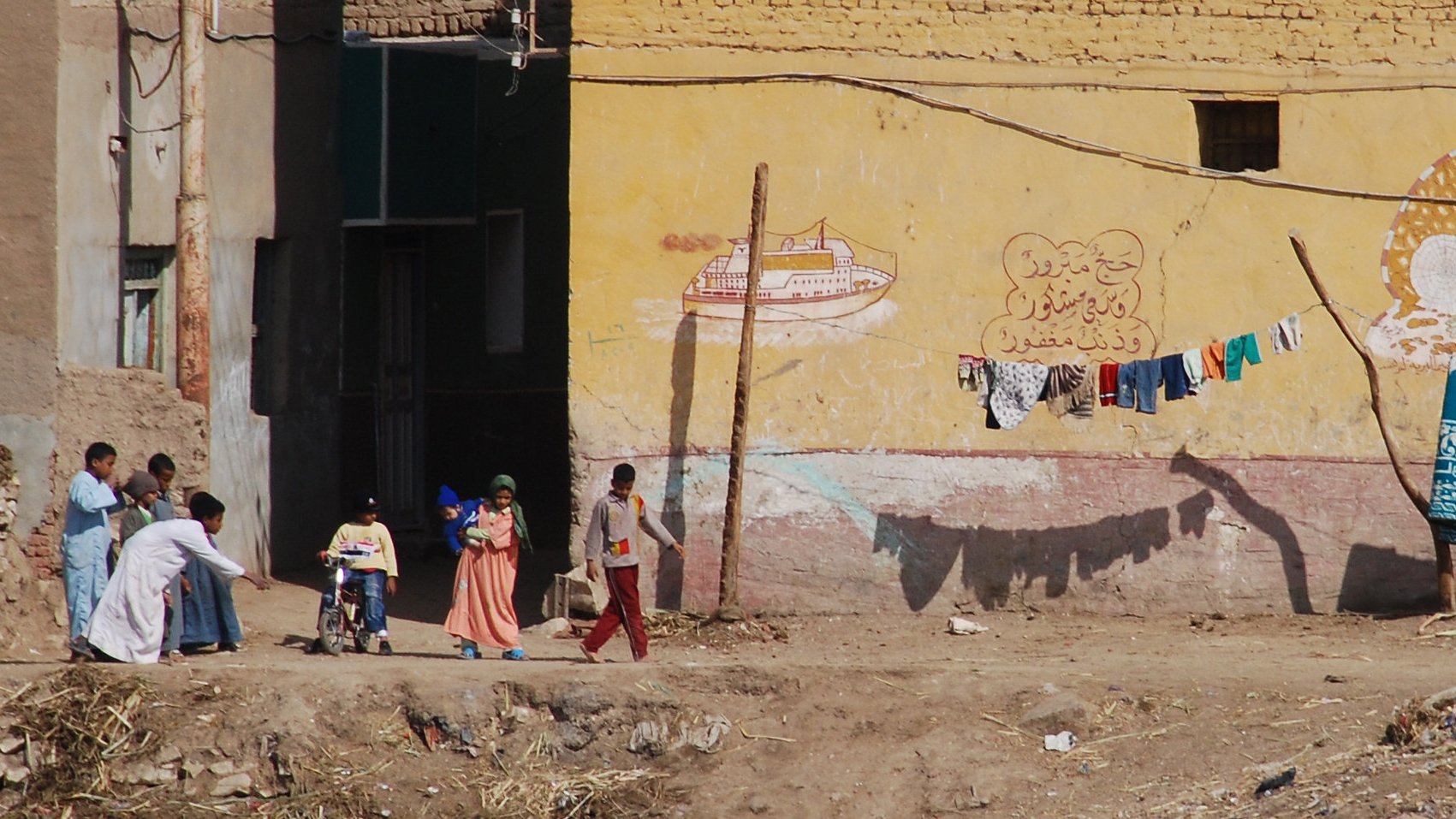 The image depicts a scene from a rural or semi-urban area, featuring a group of people interacting outside. A child in a wheelchair is being pushed forward by an adult, while two other children walk beside them. In the background, there is a building with a yellow wall that has some painted decorations or text. Above this, a clothesline displays various articles of clothing hanging to dry. The ground appears uneven, with some debris scattered around. Overall, the scene conveys everyday life in a community setting.