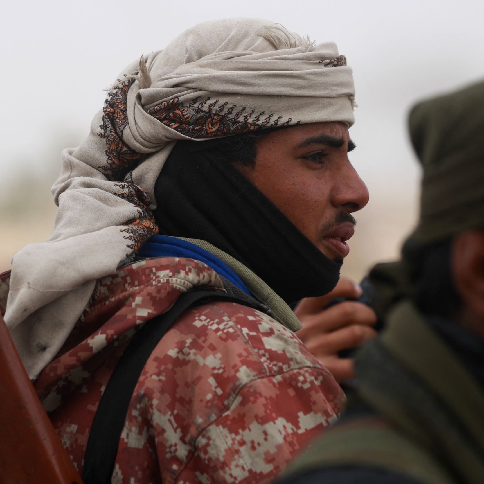 الصورة تظهر شابًا يرتدي غطاء رأس تقليدي وملابس ذات ألوان متعددة، يبدو أنه في موقف جاد أو يركز على شيء ما. يحمل سلاحًا على كتفه. خلفه يوجد أشخاص آخرون، لكن التفاصيل غير واضحة. الأجواء في الصورة قد تكون تشير إلى حالة متوترة أو سياق معين.