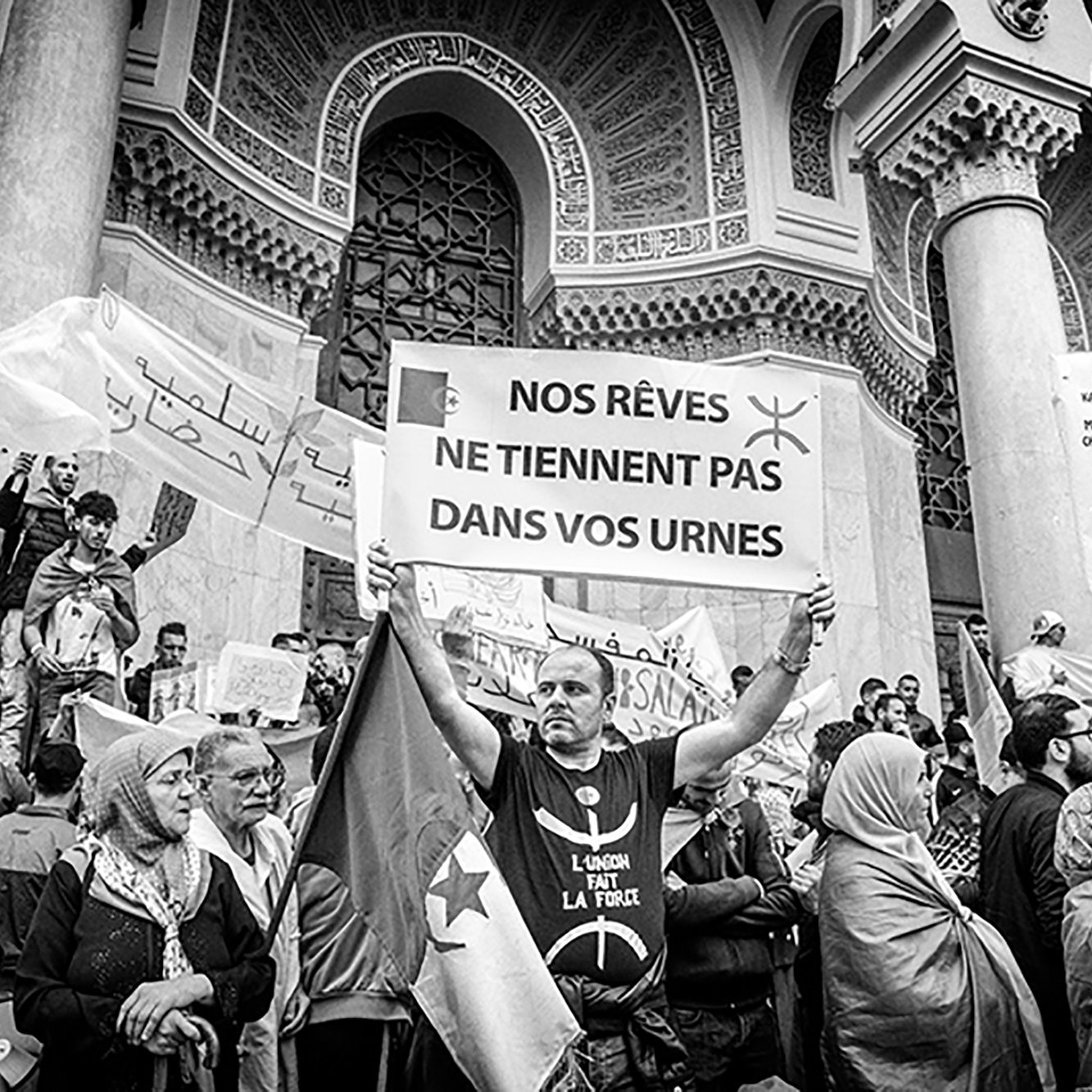 La imagen muestra una protesta o manifestación donde un grupo de personas sostiene pancartas y carteles. En el centro, un hombre sostiene un gran cartel con el mensaje "NOS RÊVES NE TIENNENT PAS DANS VOS URNES", que se traduce como "Nuestros sueños no caben en sus urnas". La escena está llena de gente, algunos con banderas y otros con rostros determinados, todos expresando descontento o reivindicaciones. El ambiente es de energía colectiva y compromiso social, y la imagen es en blanco y negro, lo que le da un tono más dramático y solemne.