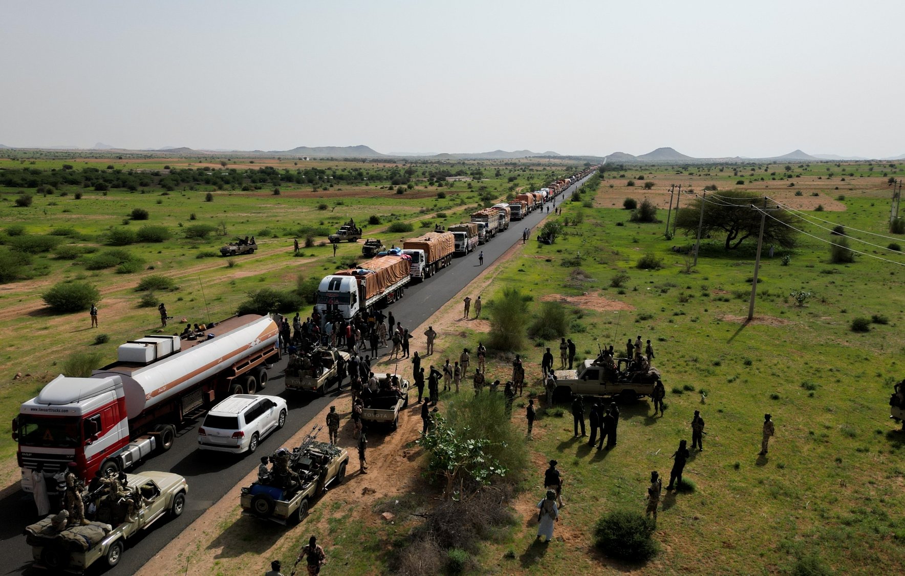 A long line of trucks on a road, with soldiers and green fields surrounding them.
