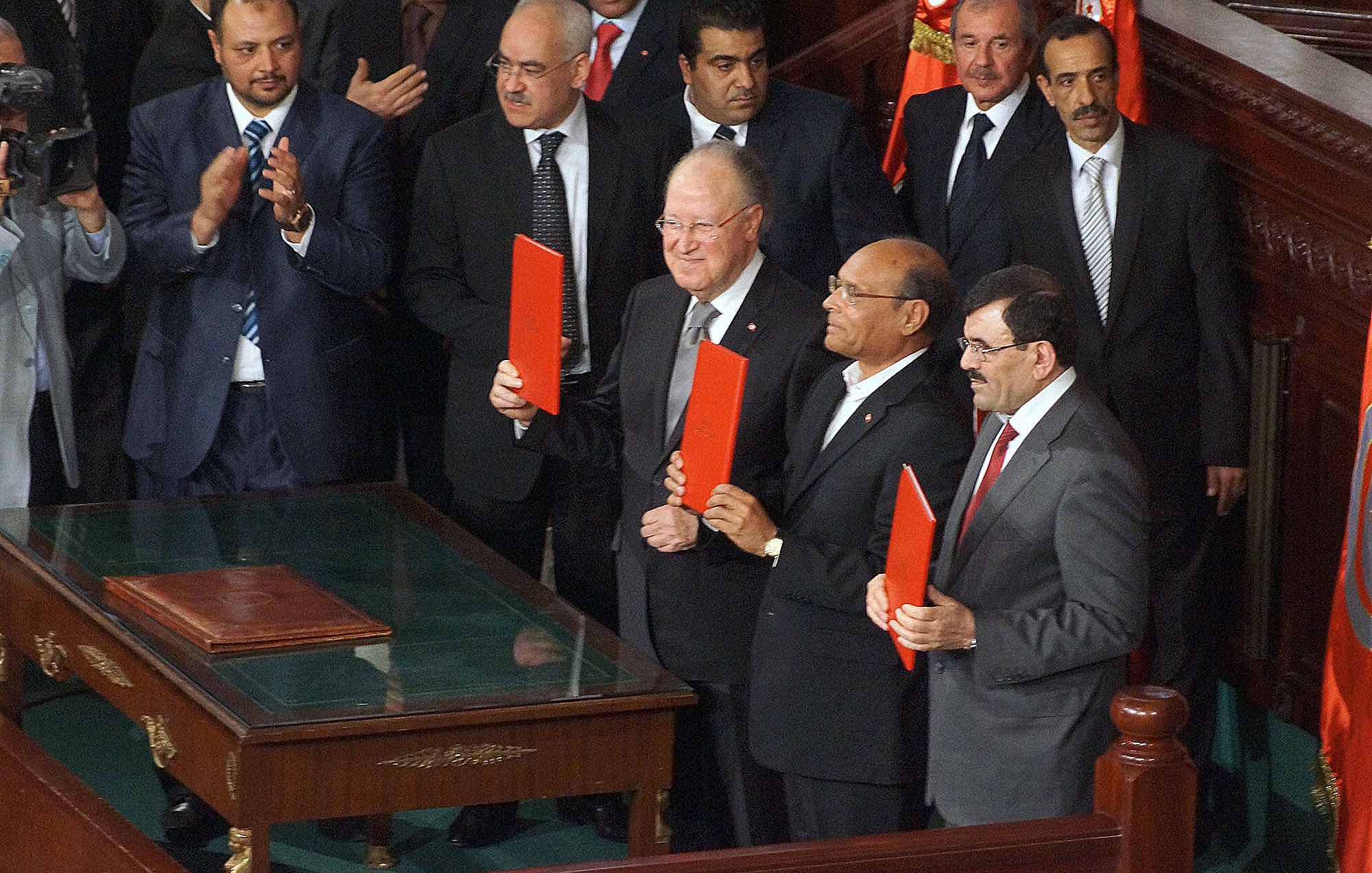 L'image montre un groupe d'hommes se tenant debout autour d'une table, tous souriants et tenant des dossiers rouges. Ils semblent être dans un cadre officiel, probablement lors d'une cérémonie ou d'une réunion importante. En arrière-plan, on peut voir d'autres personnes assises et applaudissant, ainsi que des drapeaux. L'ambiance semble solennelle et festive à la fois.