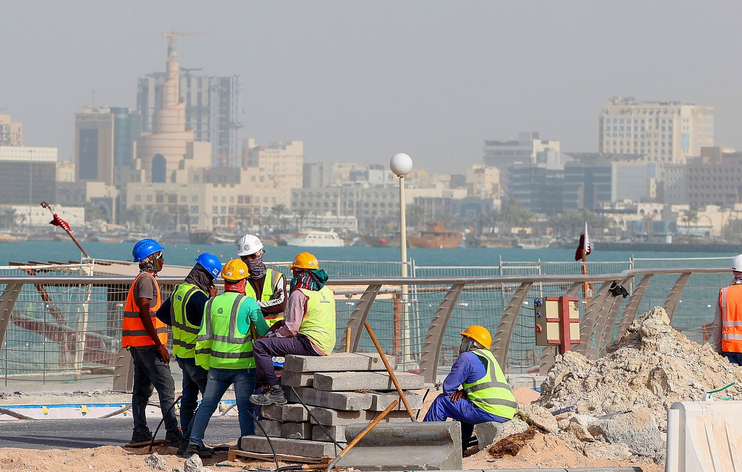 L'image montre un groupe de travailleurs sur un chantier de construction près de la mer. Ils portent des vêtements de sécurité, comme des gilets réfléchissants et des casques. En arrière-plan, on peut voir une ville avec des bâtiments modernes et des structures élevées. L'ambiance semble occupée, et le chantier est environné de matériaux de construction, avec des blocs empilés. La mer est visible au loin, ajoutant un cadre pittoresque à la scène.