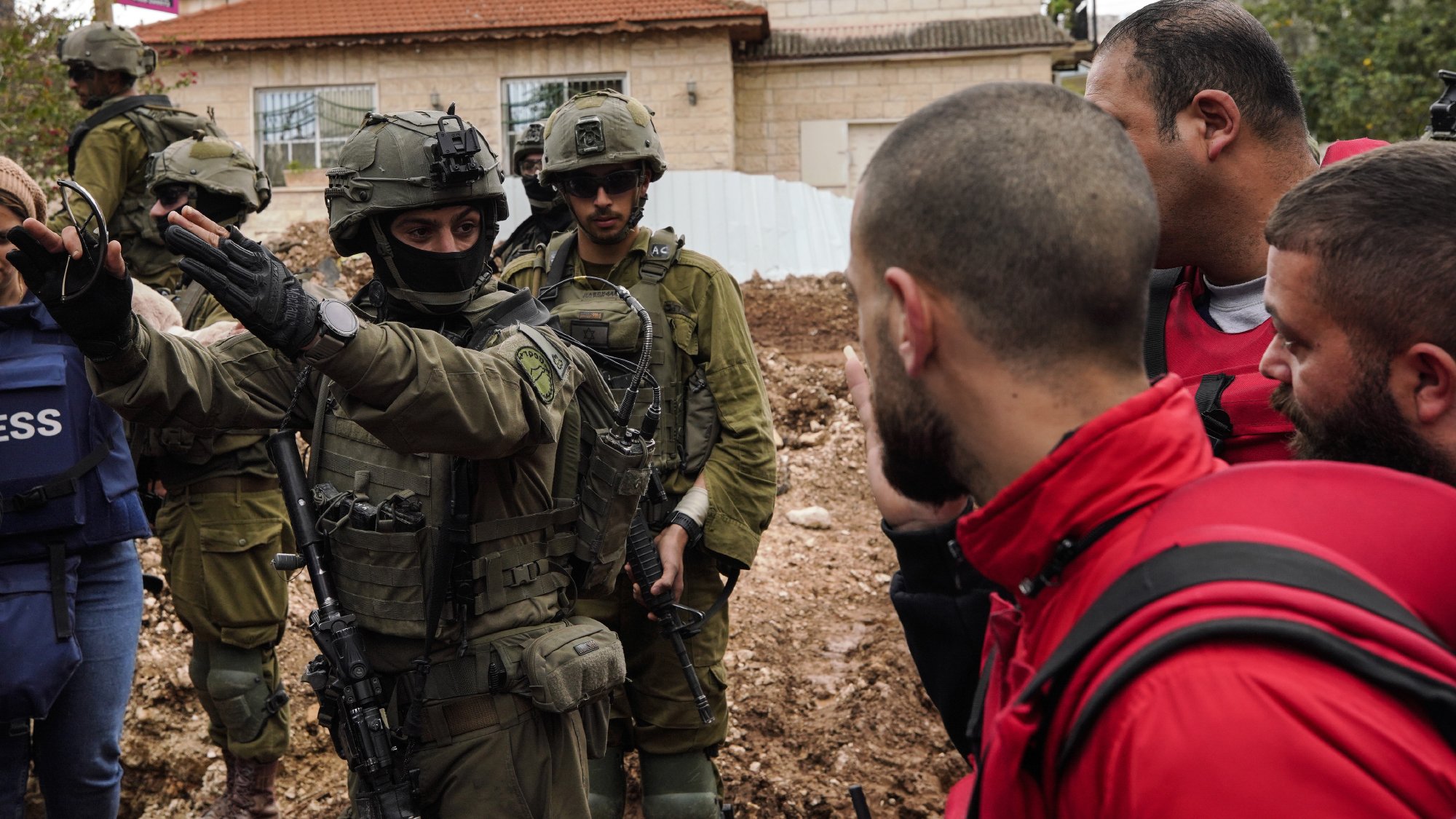 Nell'immagine ci sono diversi uomini in un contesto urbano, alcune persone indossano giacche protettive e una di loro è in uniforme militare con un'arma. Sembra esserci una discussione in corso tra le persone. Il terreno attorno a loro è fangoso e c'è un edificio sullo sfondo.