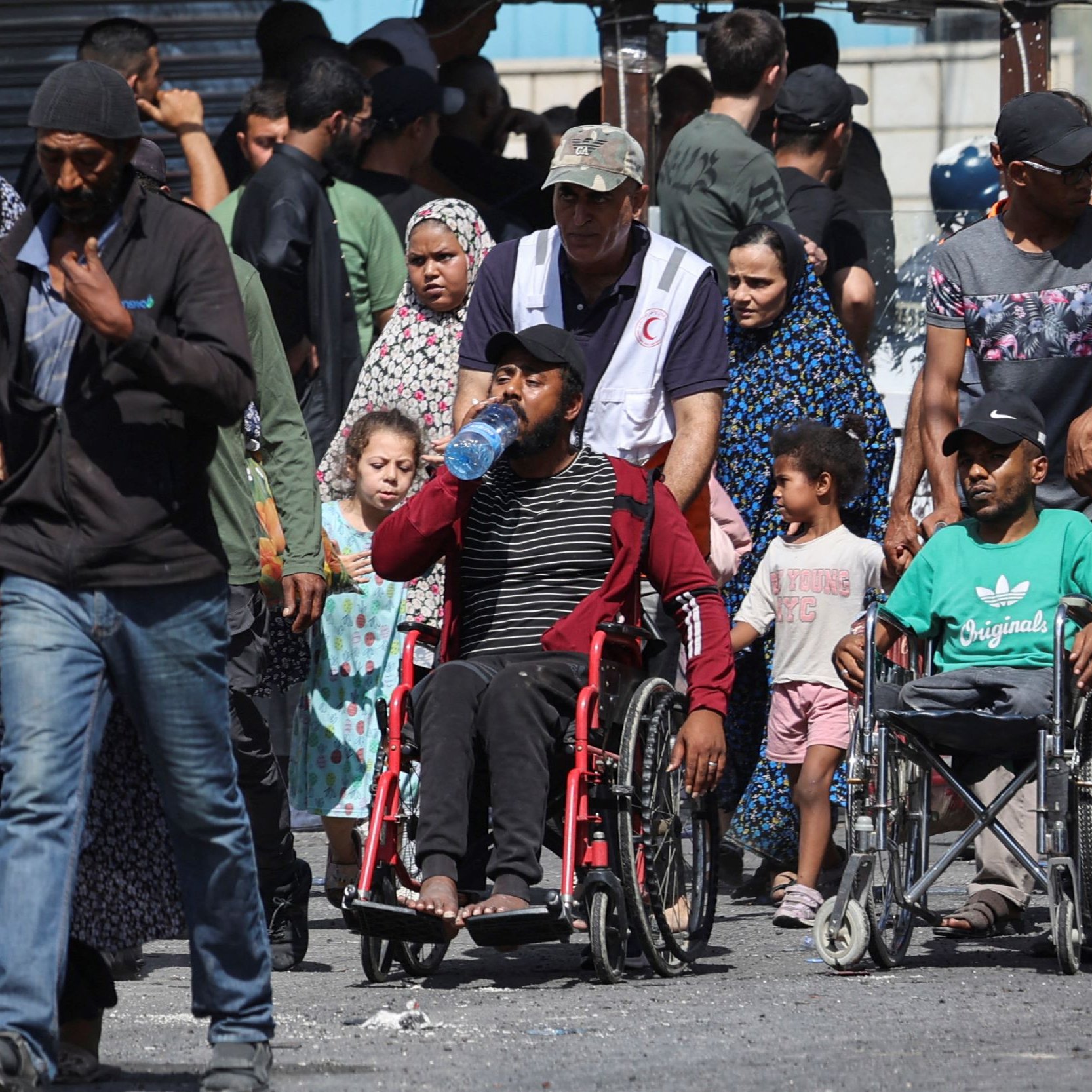 در این تصویر، گروهی از مردم در حال حرکت در یک خیابان شلوغ دیده می‌شوند. برخی از افراد در حال حمل کیف یا وسایل هستند و تعدادی از کودکان نیز در کنار بزرگ‌ترها دیده می‌شوند. فردی که روی ویلچر نشسته است به وضوح در مرکز تصویر قرار دارد و یک بطری آب در دست دارد. لباس‌های افراد متنوع است و به نظر می‌رسد که برخی از آن‌ها به طور مشخص به یک مراسم یا تجمع مربوط هستند. فضای تصویر از شلوغی و تنوع انسان‌ها پر شده است.