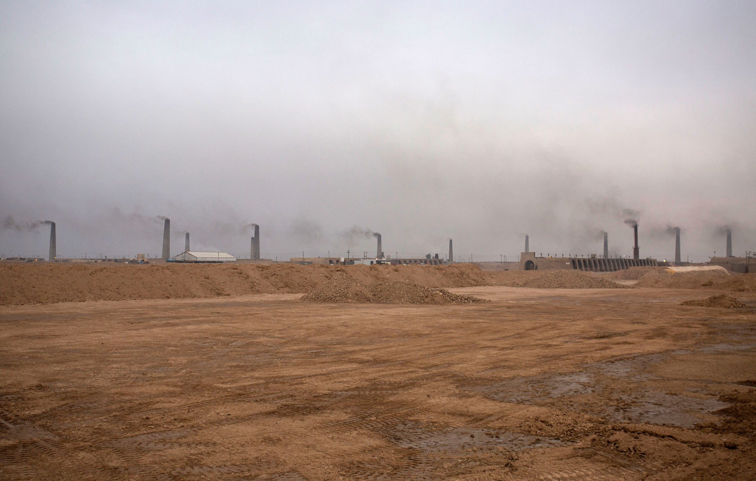 تظهر الصورة منظرًا لموقع صناعي في منطقة صحراوية، حيث يهيمن على المشهد ضباب كثيف أو سحب من الدخان تتصاعد من مداخن المنشآت. الأرض تظهر جافة وغير مستوية، مع كميات من الرمل والتربة. الأجواء تبدو رمادية وموحشة، مما يعكس تأثير النشاط الصناعي على البيئة.