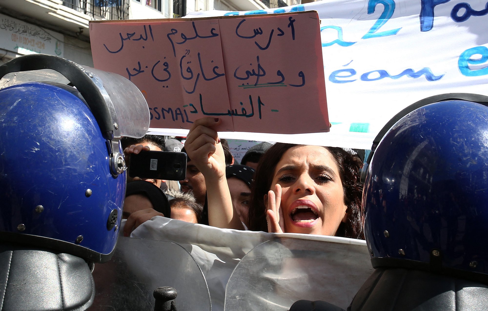 L'image montre une scène de manifestation où des personnes se rassemblent. À l'avant-plan, une femme semble exprimer des émotions vives, probablement en train de parler ou de crier, tandis qu'elle tient une pancarte. Plusieurs agents de la police, en uniforme avec des casques, se tiennent en ligne, formant un barrage. En arrière-plan, on peut apercevoir d'autres manifestants et une bannière portée par la foule. L'atmosphère semble tendue et engagée, avec une forte volonté de faire entendre des revendications.