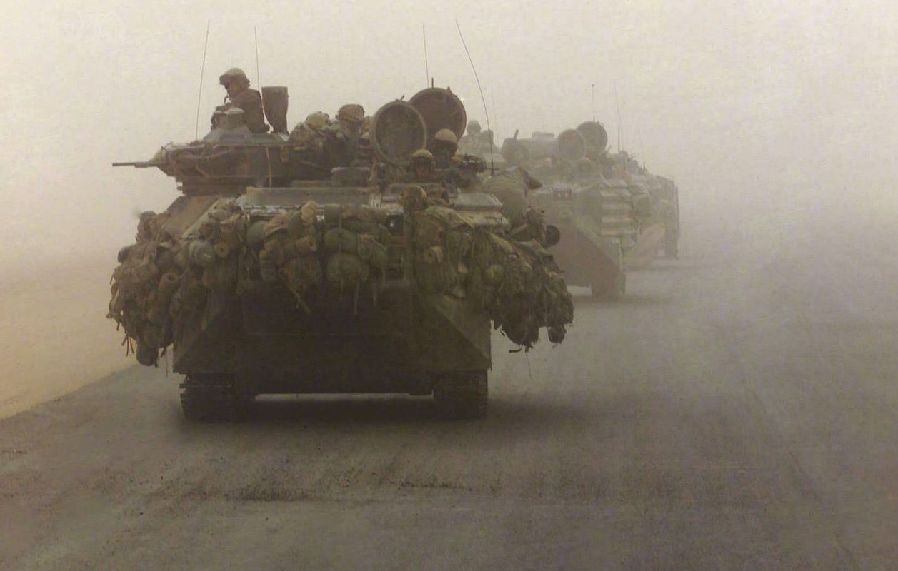 در تصویر، چندین تانک در حالی که در مه و گرد و غبار حرکت می‌کنند، به نمایش درآمده‌اند. تانک‌ها به طرز مشخصی بارگذاری شده‌اند و تجهیزات نظامی بر روی آن‌ها دیده می‌شود. محیط اطراف به شدت مه‌آلود است و دید محدود است. فضای تصویر حس تنش و حرکت در یک منطقه نظامی را منتقل می‌کند.