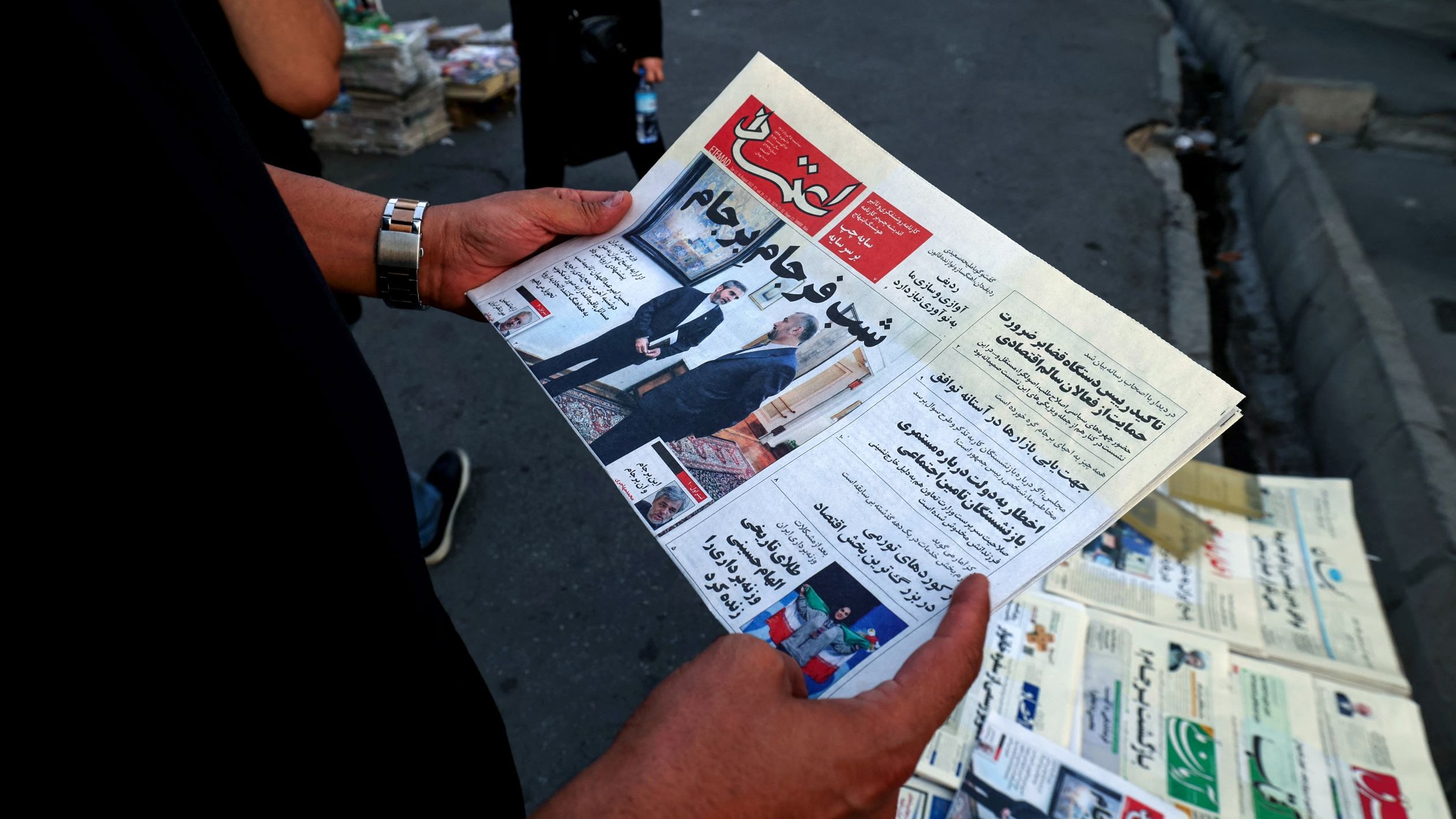 L'image montre une personne tenant un journal ouvert. Le titre est bien visible et semble relater des informations d'actualité, potentiellement politiques ou sociales. En arrière-plan, on aperçoit un kiosque à journaux avec d'autres exemplaires de journaux exposés. L'environnement semble urbain, avec des voitures garées et un sol en pavé.