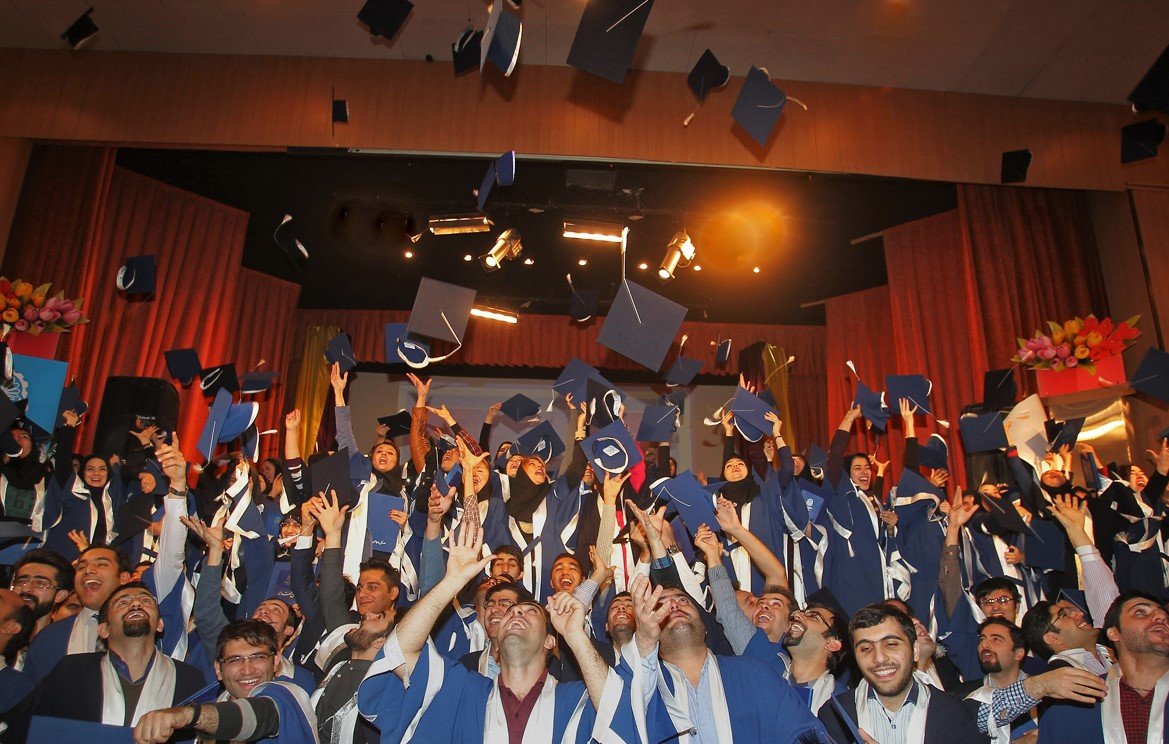 تظهر الصورة مجموعة كبيرة من الطلاب في حفل تخرج، وهم يرتدون قبعات التخرج والعباءات التقليدية. يبدو الجميع في حالة من الفرح والسعادة، حيث يرفعون قبعاتهم في الهواء احتفالاً بالإنجاز. الخلفية مليئة بالزينة، وهناك أضواء مسلطة تعزز من أجواء الحفل. تعكس الصورة روح الفخر والنجاح في هذه المناسبة المهمة في حياة الطلاب.