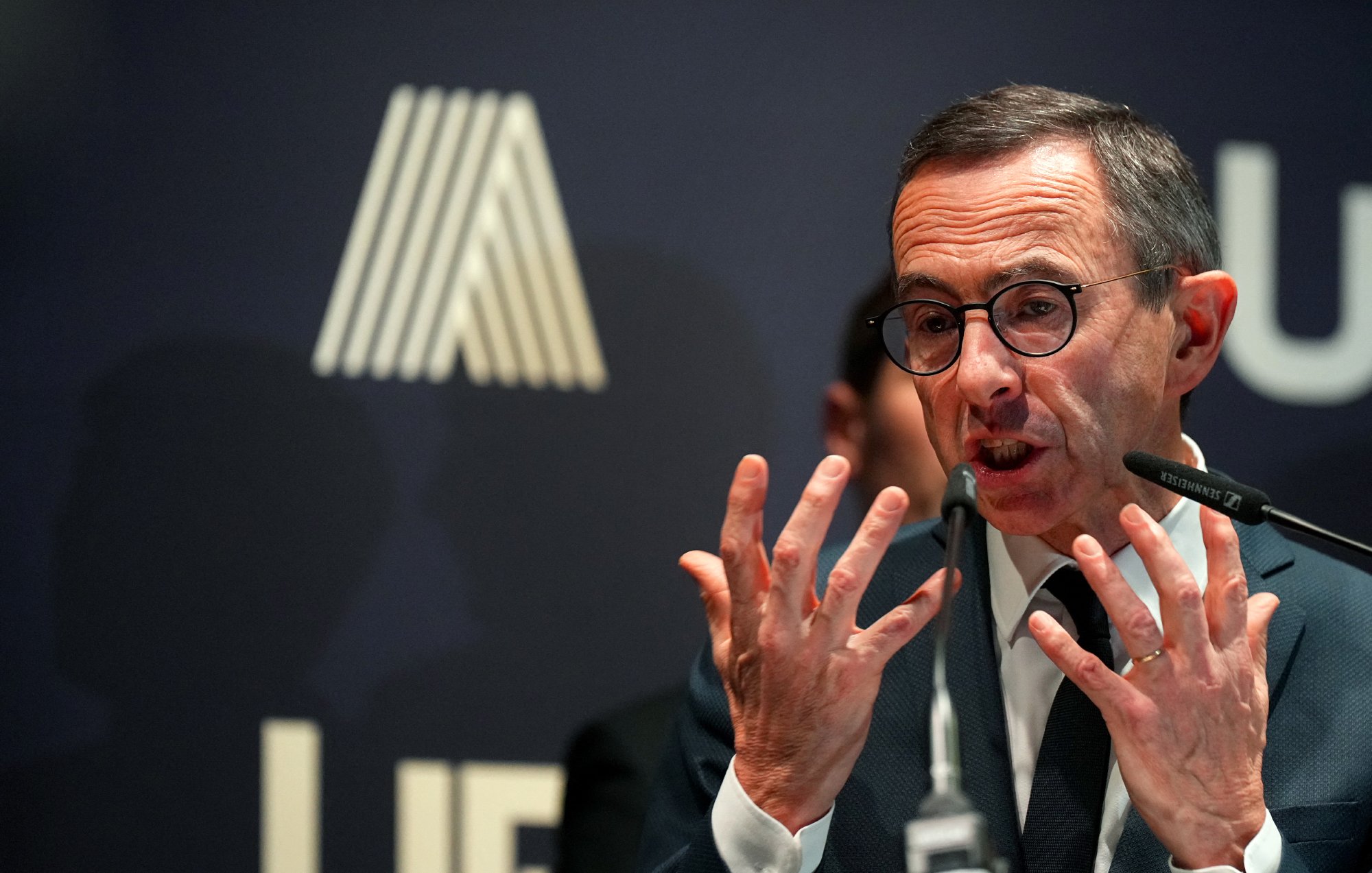 L'image montre un homme en train de parler lors d'un événement. Il porte des lunettes et a une expression animée, utilisant probablement des gestes pour renforcer son discours. En arrière-plan, on peut voir un logo qui semble être lié à une organisation, et des silhouettes floues de personnes apparaissent derrière lui. L'ambiance semble être professionnelle, suggérant que cet homme est en train de s'adresser à un public ou à des médias.