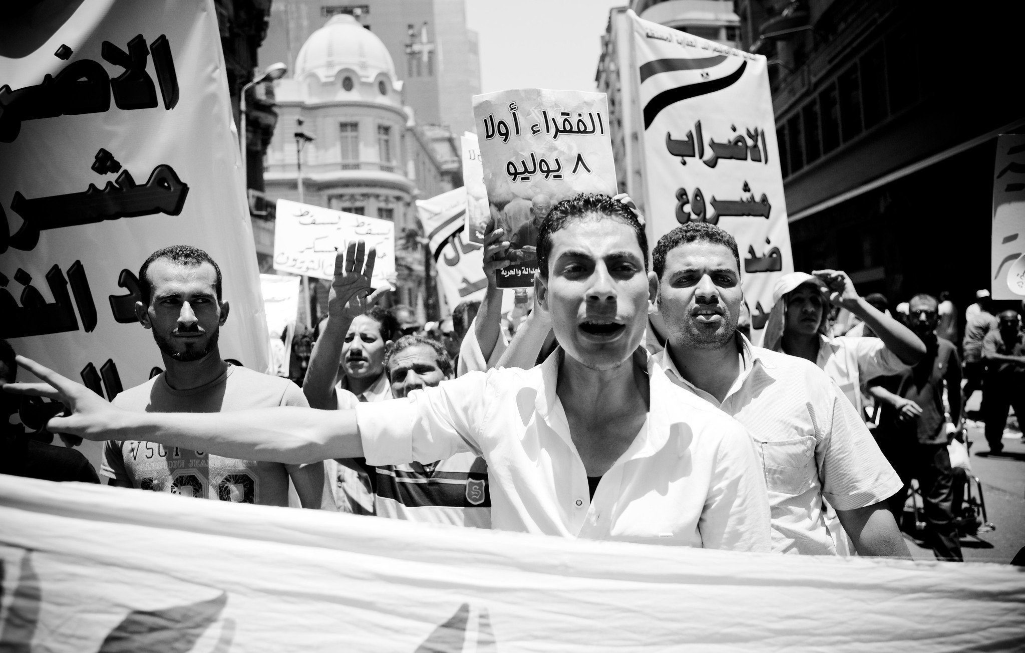 The image depicts a black and white scene of a protest or demonstration. A group of people is marching together, holding banners and signs. One person in the foreground appears to be passionately speaking or chanting, while others raise their fists or shout in the background. The signs seem to convey messages related to social or political issues, with at least one sign written in Arabic. The overall atmosphere suggests urgency and activism, capturing a moment of collective expression and dissent.