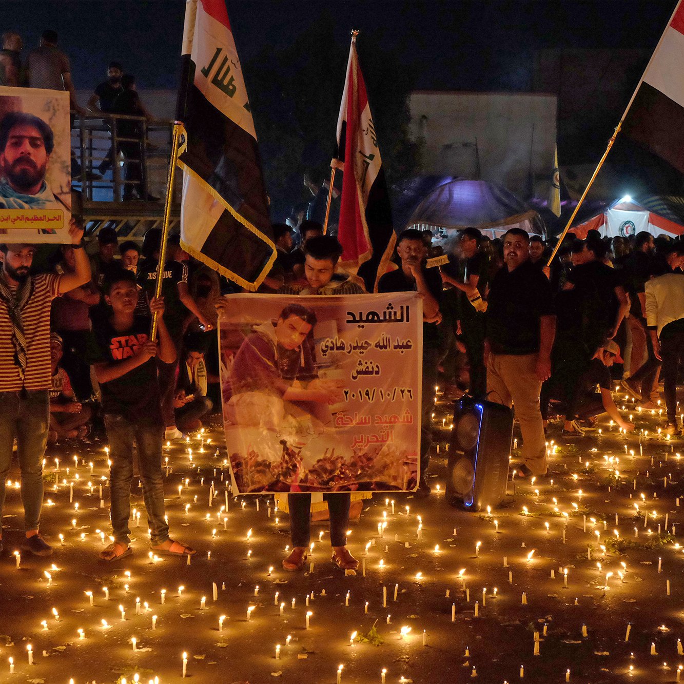 در این تصویر، جمعیتی از مردم در یک مکان بیرونی گرد هم آمده‌اند، در حال برگزاری مراسمی با شمع‌هایی روشن. برخی از افراد پرچم‌هایی در دست دارند که به نظر می‌رسد نمایانگر گروه‌های مختلفی باشد. تعدادی از شرکت‌کنندگان همچنین پوسترهایی را حمل می‌کنند که تصاویری از افراد را نشان می‌دهد. نور شمع‌ها فضای مراسم را روحانی و احساسی کرده و برجستگی آن‌ها در تاریکی شب قابل توجه است. این تجمع ممکن است به منظور یادبود یا گرامیداشت افرادی خاص باشد.