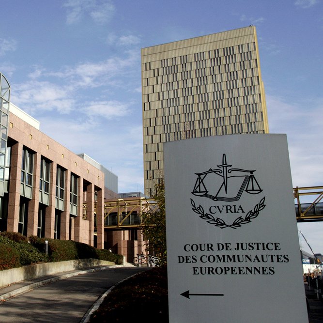 L'image montre le bâtiment de la Cour de justice des communautés européennes, situé à Luxembourg. On peut y voir une architecture moderne avec des éléments en verre et en pierre. À l'avant-plan, il y a un panneau indiquant le nom de la cour, accompagné de symboles de la justice. L'ensemble est entouré d'un aménagement paysager, avec des arbustes et un chemin qui mène à l'entrée. Le ciel est partiellement nuageux, ajoutant une ambiance calme à la scène.