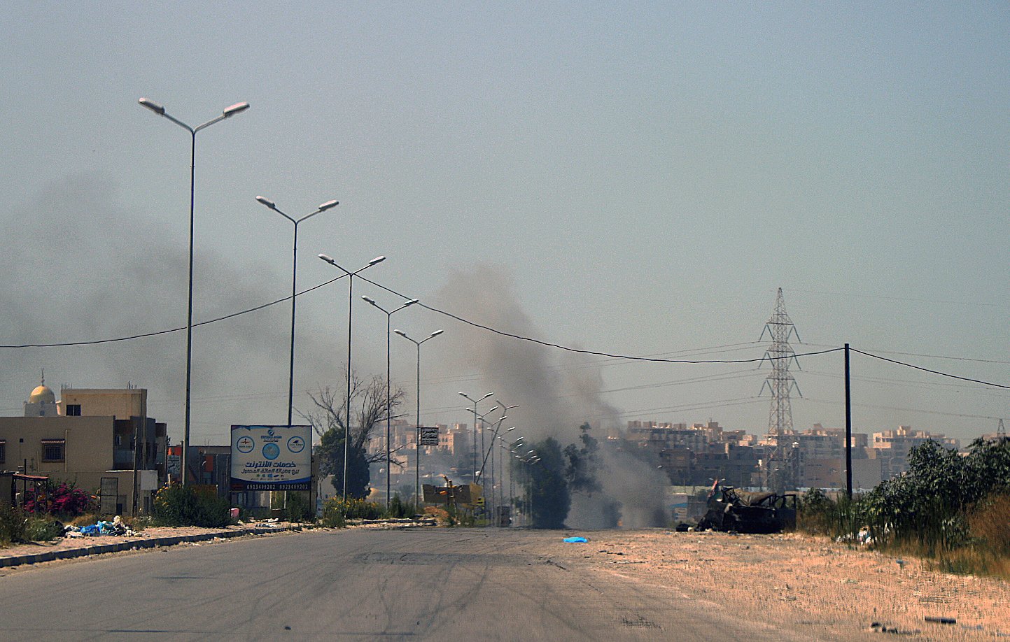 L'image montre une scène de route apparemment désertée, avec quelques bâtiments sur les côtés. On peut apercevoir de la fumée s'élevant au loin, indiquant probablement un incendie ou une explosion. Des panneaux de signalisation sont visibles, ainsi que des lampadaires le long de la route. L'atmosphère semble chargée et la visibilité est atténuée par la fumée.