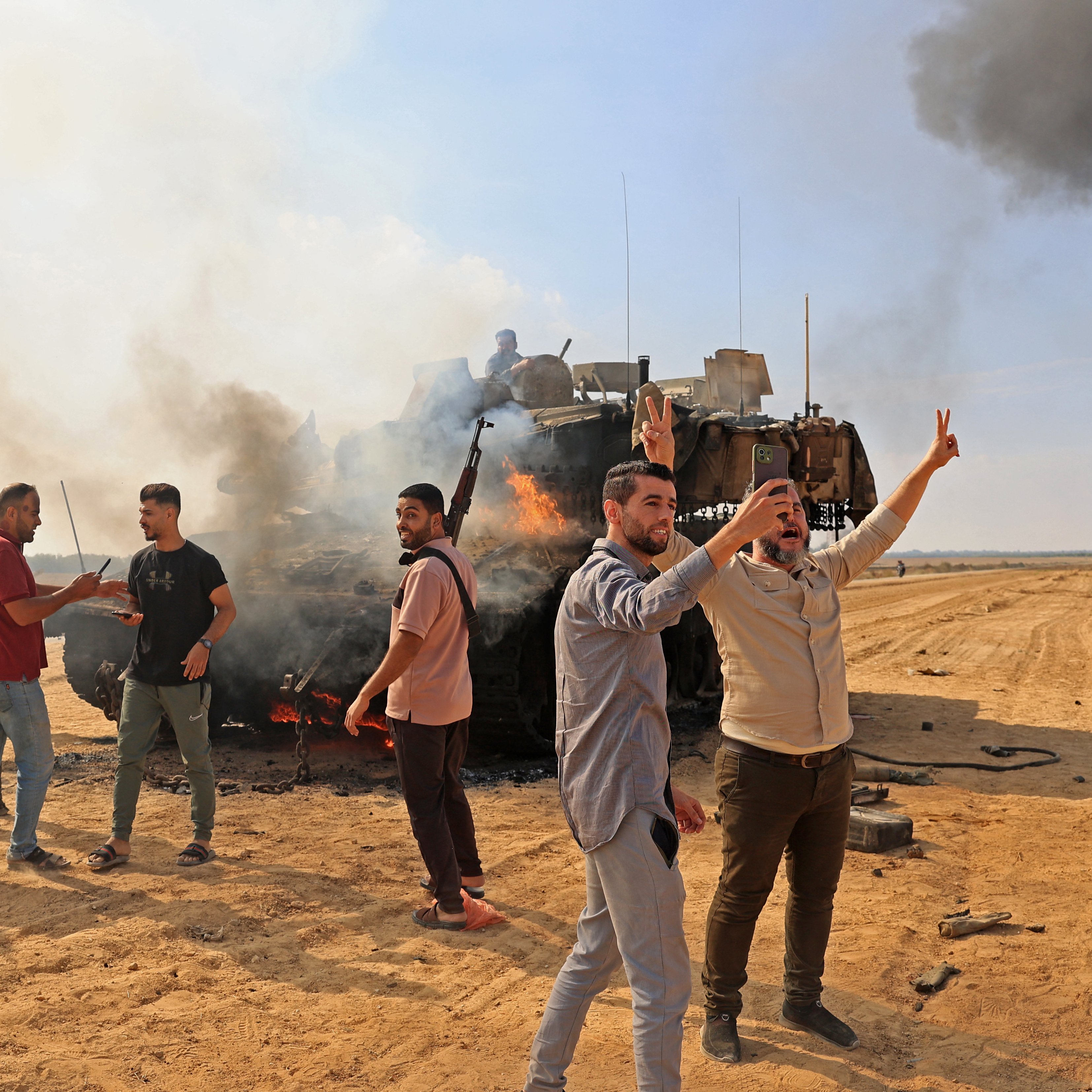 تظهر الصورة مجموعة من الأشخاص في منطقة صحراوية تتصاعد فيها الدخان من عربة مدرعة مدمَّرة. بعض الأشخاص يرفعون أيديهم في إشارة النصر أو الاحتفاء، بينما يبدو آخرون مشغولين بالتفاعل مع بعضهم البعض. الأجواء تعكس حالة من التوتر، مع وجود عمود من الدخان الأسود في الخلفية. السماء زرقاء ولكن مشوشة بسبب الدخان.