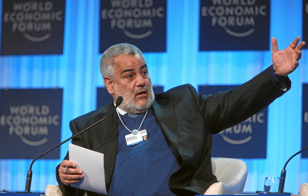 L'image montre un homme en train de parler lors d'un événement, probablement une conférence ou un forum. Il porte un costume noir avec un pull bleu en dessous. Il semble être engagé dans une discussion, levant le bras pour souligner un point. En arrière-plan, on peut voir des panneaux portant le logo du Forum économique mondial. L'ambiance paraît professionnelle et dynamique.