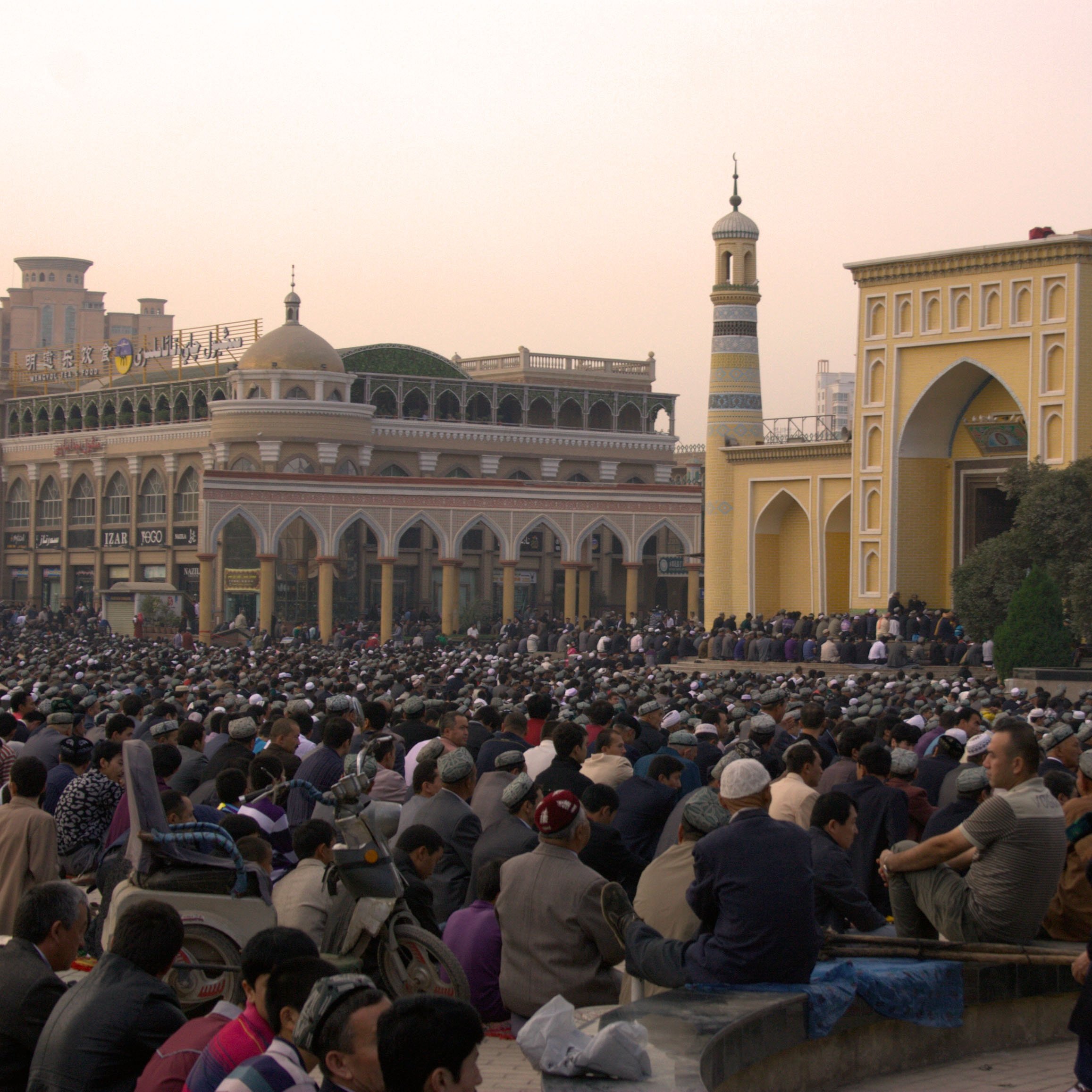 L'image montre une grande foule de personnes rassemblées dans une zone en plein air, probablement pour un service religieux. À l'arrière-plan, on aperçoit des bâtiments architecturaux impressionnants, avec des minarets et des ornements qui évoquent une ambiance culturelle riche. Les gens sont principalement assis, attentifs à ce qui se passe, créant une atmosphère de commémoration ou de prière collective. La lumière du soir donne une teinte chaleureuse à la scène.