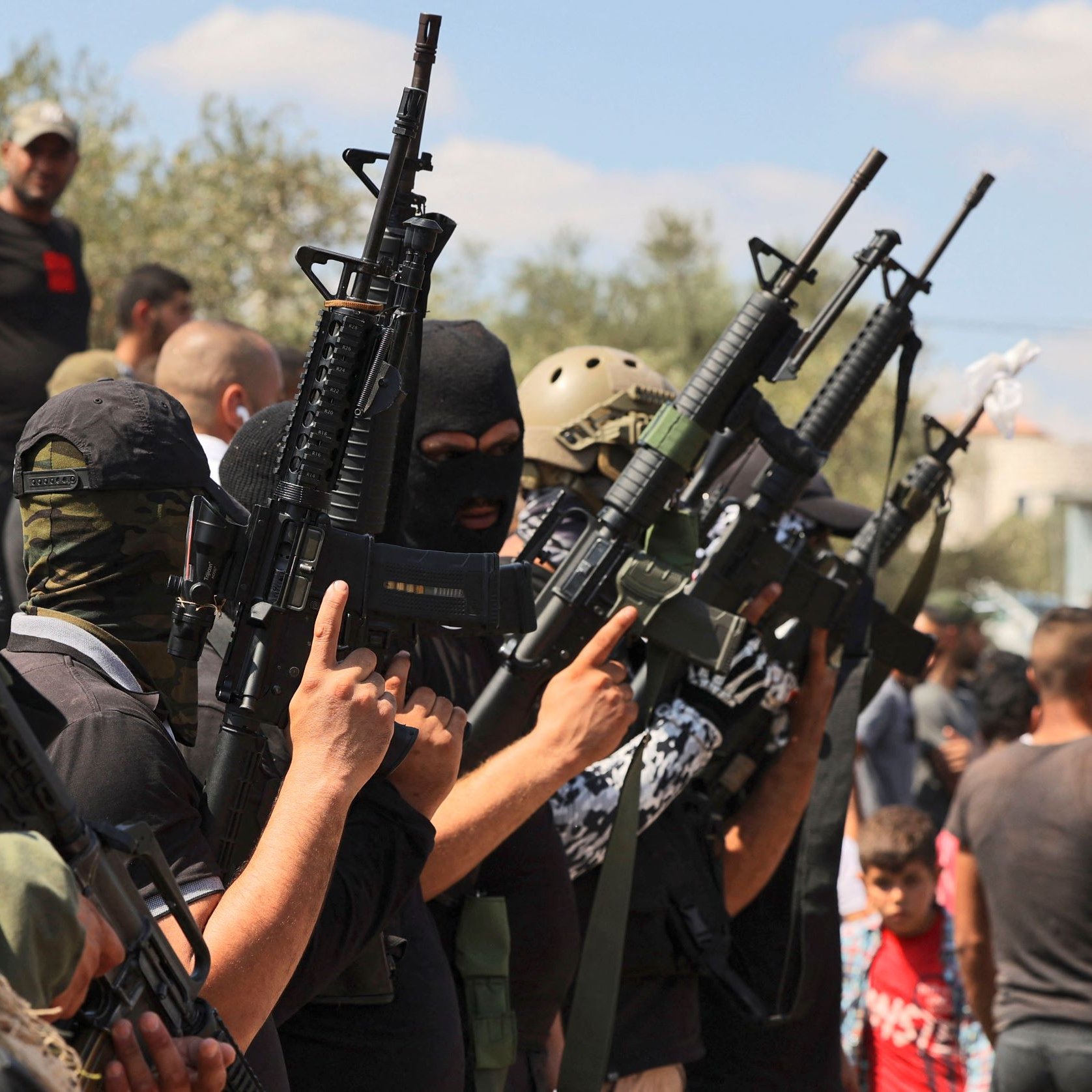 در این تصویر، گروهی از مردان با ماسک‌های سفید و مشکی که صورت‌هایشان را می‌پوشاندند، در حال ایستادن هستند. آن‌ها سلاح‌های اتوماتیک در دست دارند و لباس‌های نظامی به تن دارند. در پس‌زمینه، افراد دیگری به وضوح دیده می‌شوند که به این گروه نگاه می‌کنند. فضا به نظر حالت نظامی و جدی دارد.