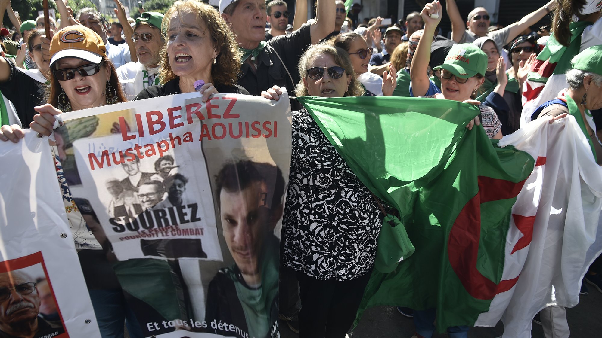 L'image montre une manifestation où des personnes sont rassemblées, exprimant des revendications. On peut voir des individus brandissant des pancartes et des drapeaux, notamment un drapeau algérien. Les manifestants semblent mobilisés et engagés, scandant des slogans pour la libération de Mustapha Aouissi. L'atmosphère semble dynamique et pleine d'énergie, avec une forte présence de diversité parmi les participants.