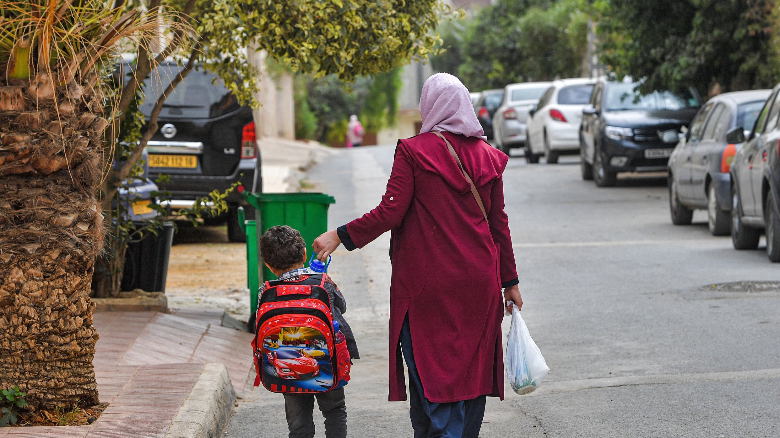 The image shows a woman walking down a street with a young child. The woman is wearing a long maroon coat and a headscarf, while the child has a colorful backpack with a cartoon character design. They are walking hand in hand along a road lined with parked cars and greenery on the sides. The atmosphere appears casual and everyday, suggesting they are possibly returning from school or heading to a local destination.