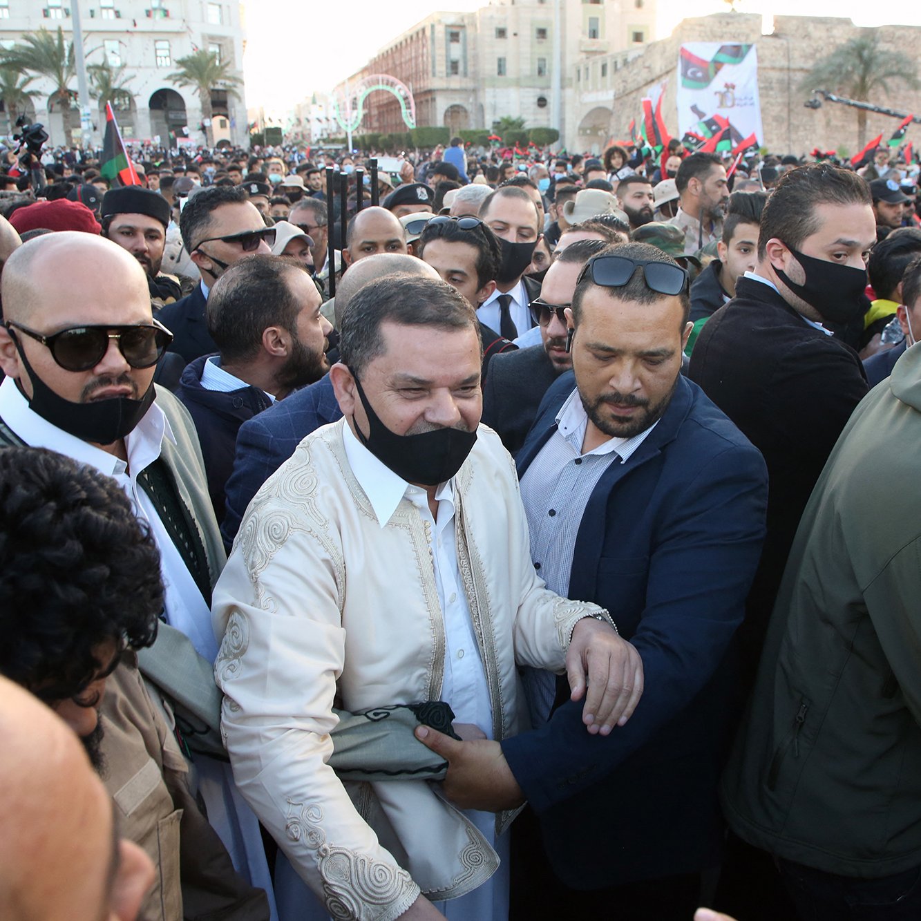 The image depicts a crowd of people gathered at an outdoor event, likely a political or social gathering. In the foreground, a man wearing a white traditional outfit is surrounded by a group of security personnel or supporters. Many individuals in the crowd are wearing black face masks. The background features various flags and buildings, indicating a festive or significant atmosphere. The scene appears to be lively, with a sense of excitement among the gathered individuals.