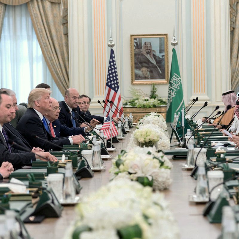 L'image montre une réunion formelle dans une grande salle avec une longue table. D'un côté, plusieurs représentants américains, dont on peut distinguer deux hommes en costume, ainsi qu'une personne qui semble être le président. En face d'eux, des dignitaires saoudiens, également en costume traditionnel. La salle est ornée de drapeaux américain et saoudien, et des arrangements floraux décoraient la table. L'atmosphère semble sérieuse et collaborative.