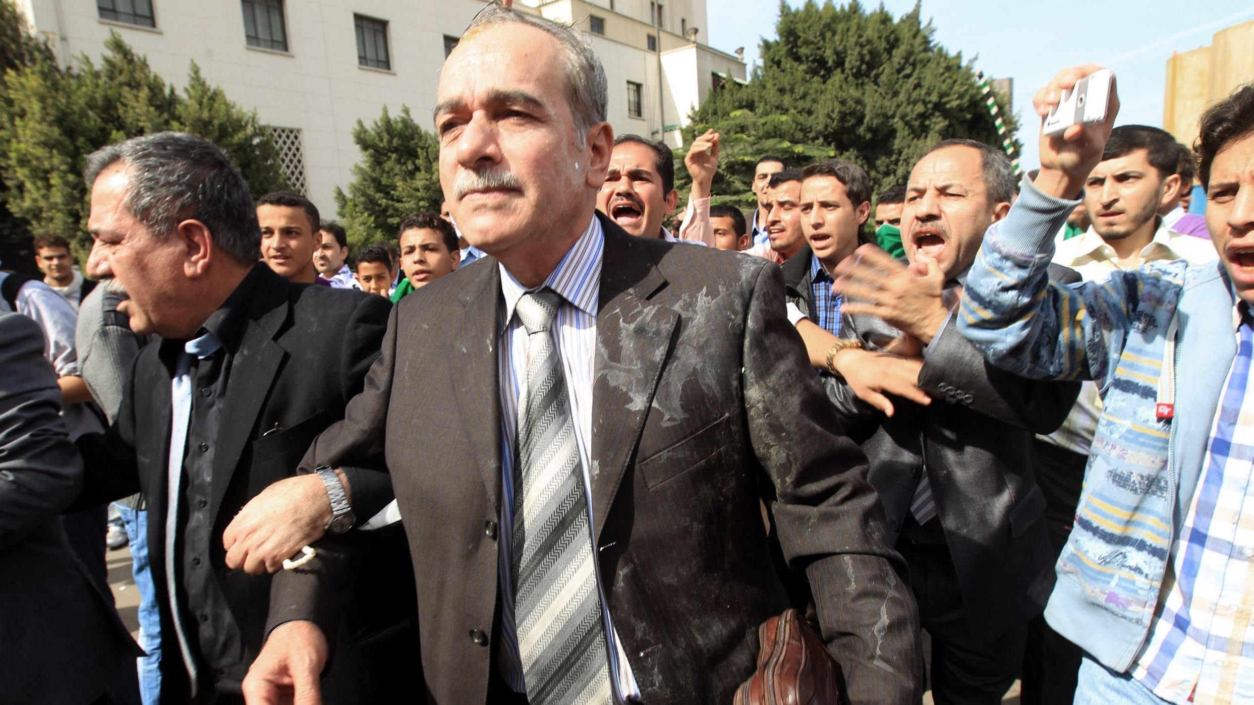 تظهر الصورة مجموعة من الأشخاص في مكان عام، حيث يبدو أن هناك تفاعل قوي ومثير. في المقدمة، يوجد رجل يرتدي بدلة رسمية ويظهر على وجهه تعبير جدّي. يحيط به عدد من الأشخاص الآخرين الذين يبدو أنهم متحمسون أو منفعلون، مما يوحي بوجود حدث مهم أو احتجاج. الخلفية تضم مجموعة من الأشجار والمباني، مما يعطي انطباعًا بأن المكان هو منطقة حضارية. الجو العام في الصورة يبدو ديناميكيًا ومليئًا بالطاقة.