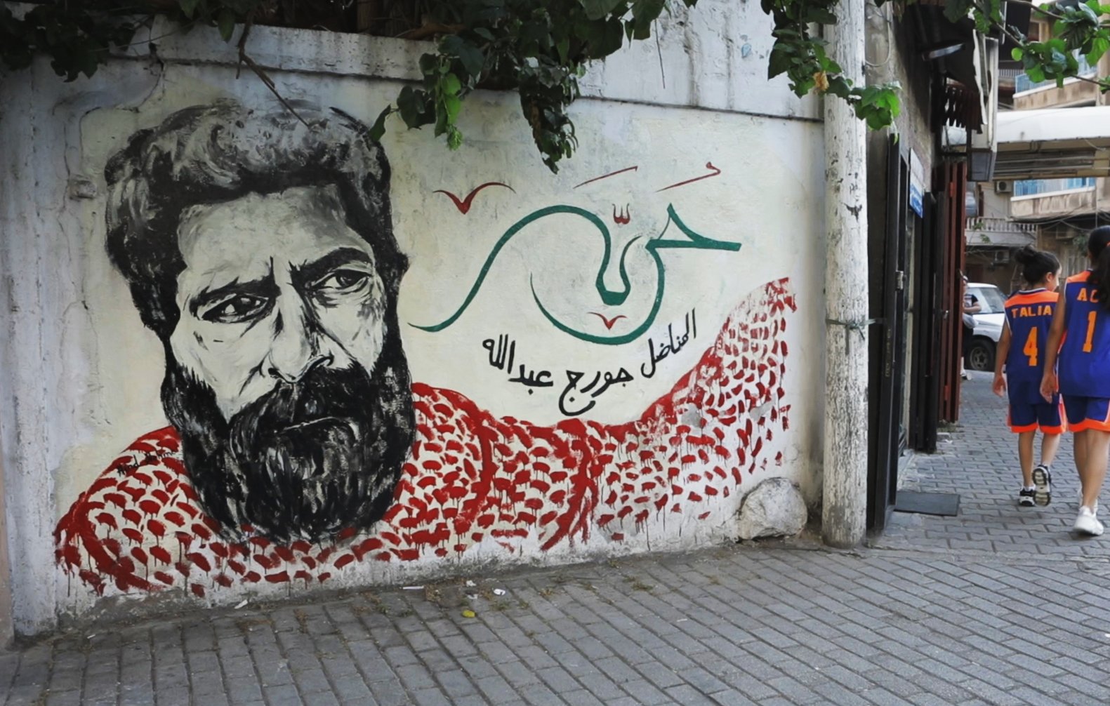L'image montre une fresque murale d'un homme avec une barbe, probablement un portrait stylisé. Il porte un keffieh rouge et blanc, et le fond est orné de motifs et de couleurs vives. À côté, deux personnes marchent sur un trottoir, portant des vêtements de sport. L'environnement semble urbain, avec des bâtiments en arrière-plan et de la végétation. Les éléments de la fresque et la scène de rue créent une atmosphère vivante et culturelle.