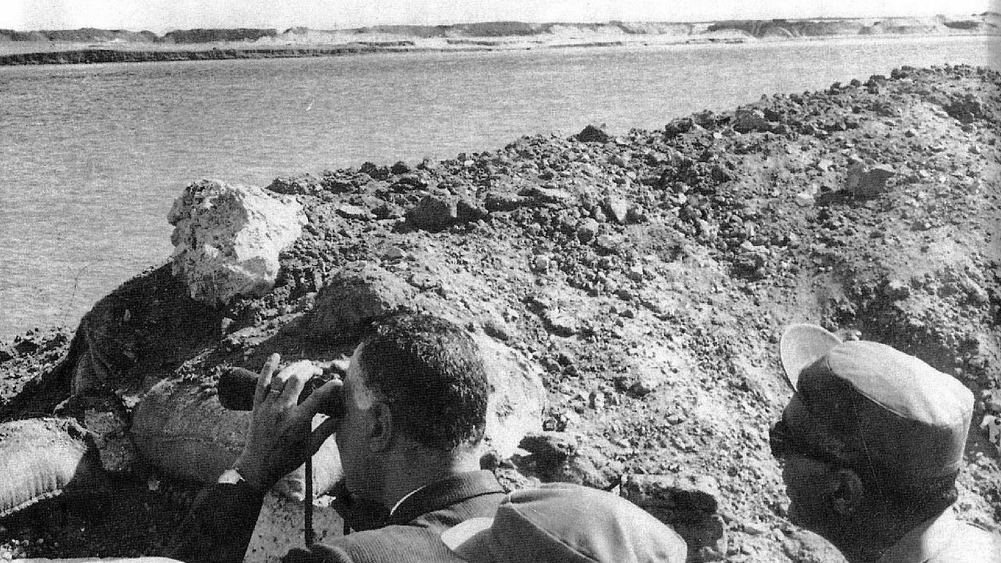 تظهر في الصورة مجموعة من الأشخاص يقفون على حافة منطقة مائية، يراقبون شيئًا ما باستخدام المنظار. يبدو أن الشخص الموجود في المقدمة مشغول بالنظر من خلال المنظار، بينما يراقبه الآخرون من حوله. الخلفية تتكون من تربة وعناصر طبيعية، مما يوحي بأن الصورة قد التُقطت في موقع استراتيجي أو أثناء زيارة ميدانية.