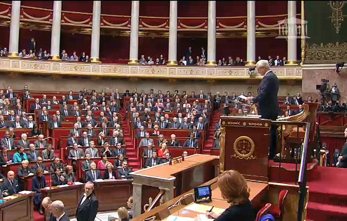 L'image montre une scène à l'Assemblée nationale, un lieu de débat politique en France. On y voit un grand amphithéâtre avec de nombreux sièges, occupés par des députés. Au premier plan, un homme se tient debout devant un pupitre, probablement en train de faire un discours ou de présenter une proposition. Les députés semblent attentifs et sont disposés en plusieurs rangées, tandis que certains sont assis à des tables sur le côté. L'architecture est typiquement institutionnelle, avec des éléments ornementaux et une atmosphère solennelle.