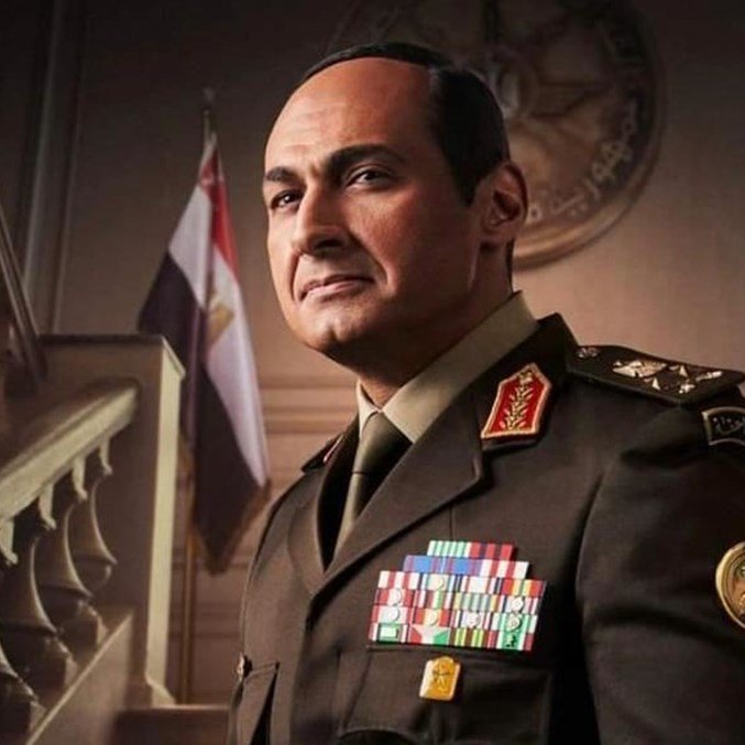 يظهر في الصورة رجل يرتدي زيًا عسكريًا يحمل العديد من الأوسمة والرتب. يبدو أنه يتخذ وضعًا رسميًا وجادًا، مع خلفية تتضمن علمين. تعكس تعابير وجهه الثقة والسلطة، كما أن التصميم العام للصورة يوحي بأجواء عسكرية أو حكومية.