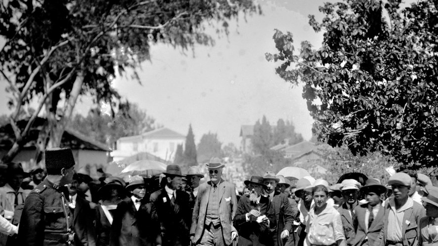 در این تصویر، گروهی از مردم در یک فضای باز دیده می‌شوند. بخشی از آنها چترهایی در دست دارند و ظاهراً در حال تماشای یک رویداد یا شخصیت مهم هستند. در وسط تصویر، مردی با عصا و لباس رسمی در حال حرکت است. اطراف او جمعیتی از مردان و زنان ایستاده‌اند که همگی لباس‌های رسمی و مرتب به تن دارند. در سمت چپ، فردی در لباس نظامی دیده می‌شود که به نظر می‌رسد به عنوان نگهبان یا محافظ عمل می‌کند. فضای عمومی و هوای آفتابی نشان‌دهنده یک روز شلوغ و پرحادثه است.