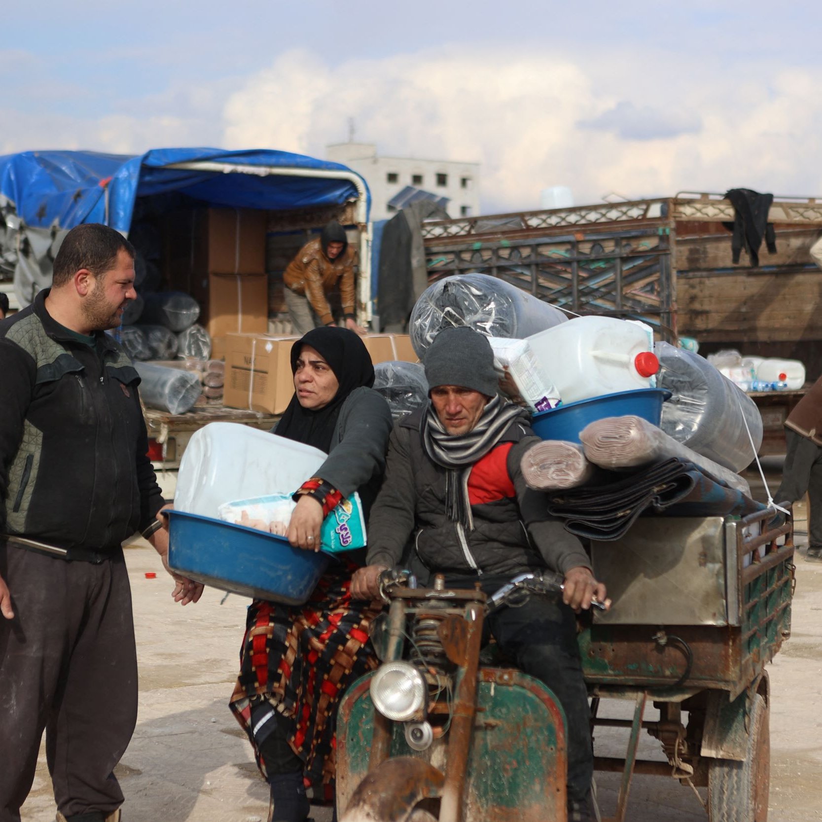The image shows a busy scene where people are engaged in various activities, likely in a market or a distribution area. In the foreground, a man and a woman are interacting next to a small vehicle loaded with large containers, possibly for water or other supplies. The woman is dressed in traditional clothing and appears to be holding a tray, while the man seems to be dressed in casual attire. In the background, several people are seen unloading boxes and organizing goods, with a truck and some tents visible. The setting has a somewhat informal atmosphere, suggestive of a community effort related to aid or distribution.