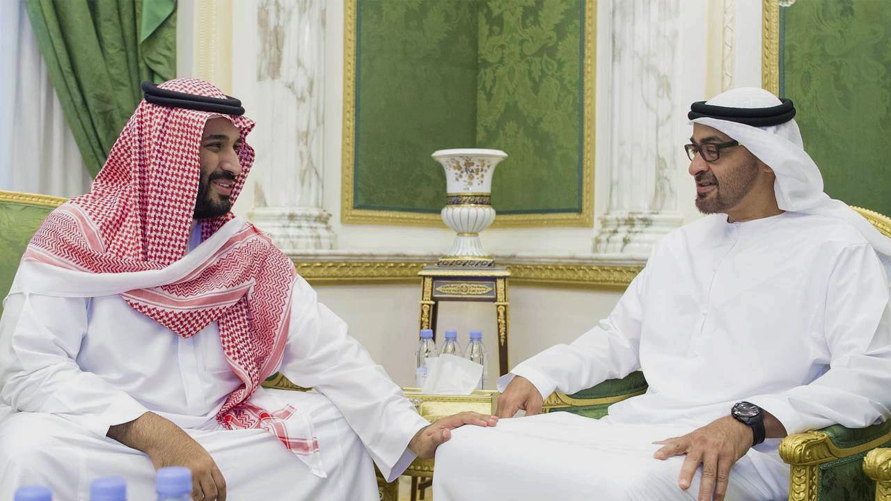 تظهر في الصورة اثنان من الرجال يجلسان معًا في غرفة مزينة بطريقة فاخرة. يرتدي الأول الزي السعودي التقليدي مع الشماغ الأحمر والأبيض، بينما يرتدي الثاني الزي الإماراتي التقليدي. كلاهما يبدو سعيدًا، ويتبادل الحديث في أجواء ودية. الخلفية تتضمن تفاصيل خضراء وذهبية، مما يضيف جوًا من الرقي والفخامة. على الطاولة أمامهم زجاجات ماء، مما يشير إلى اجتماع أو مناسبة اجتماعية.