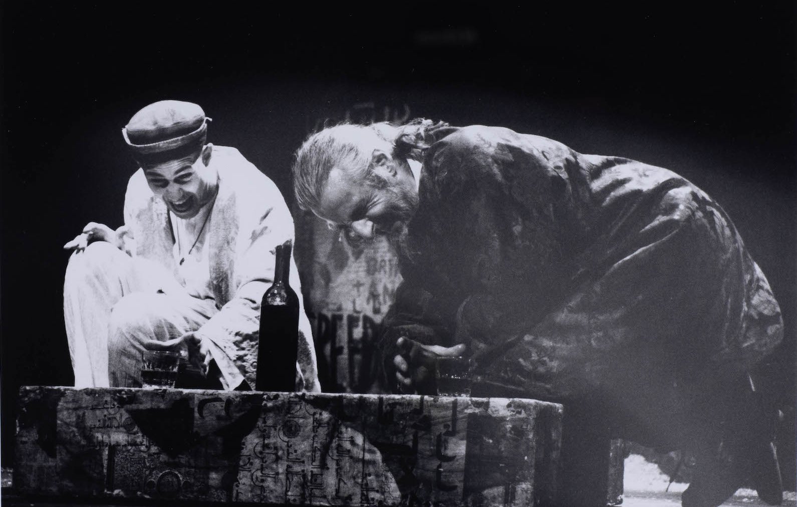 Deux hommes en scène, riant, près d'une bouteille sur une caisse au fond sombre.