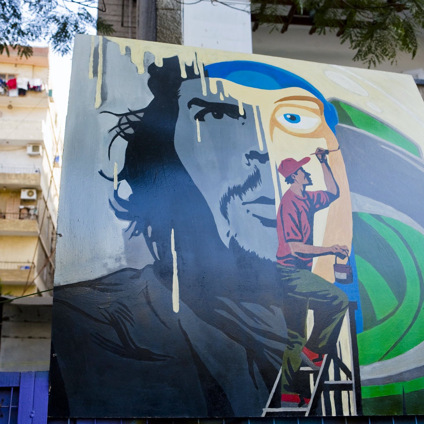 L'image montre une fresque murale colorée. À gauche, il y a le visage d'un homme avec une expression intense, tandis qu'à droite, un artiste est représenté en train de peindre. L'artiste est vêtu d'un pantalon rouge et d'un t-shirt, et il est sur une échelle, concentré sur son œuvre. En arrière-plan, on aperçoit des bâtiments avec des balcons et du linge suspendu. Les couleurs de la fresque sont vives, avec des nuances de bleu et de vert qui contrastent avec les éléments plus sombres du visage.