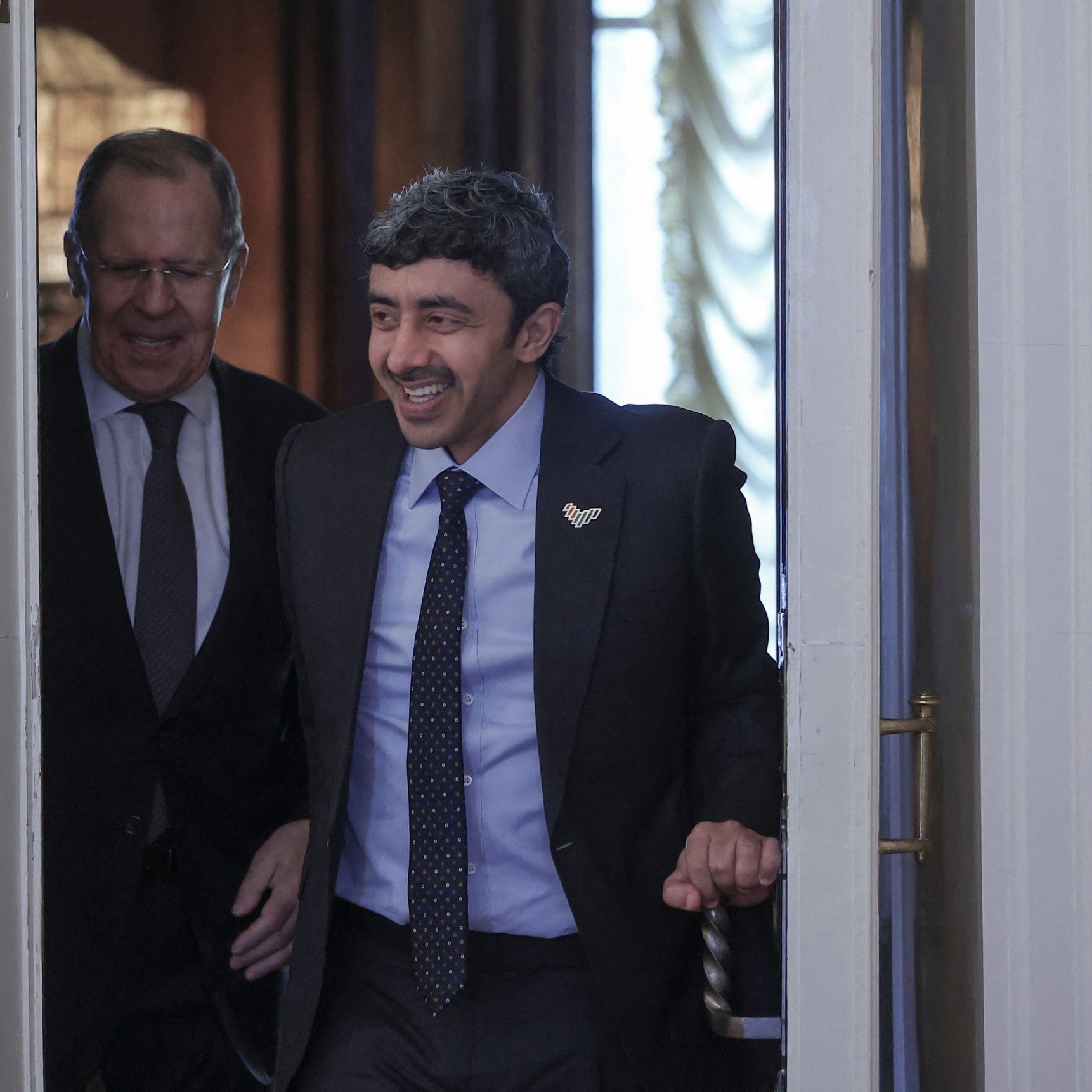 La imagen muestra a dos hombres entrando en una sala. Uno de ellos tiene una sonrisa amplia y está vestido con un traje oscuro y una corbata con un patrón. El otro hombre parece estar sonriendo también y lleva unas gafas. La decoración del fondo sugiere un ambiente formal y elegante, con luces suaves y detalles arquitectónicos. Ambos parecen estar en un momento de cordialidad y alegra.