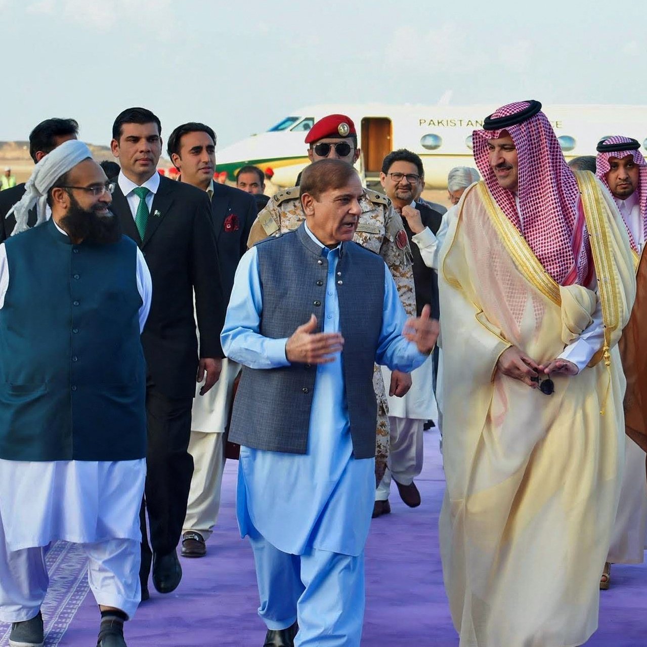 در این تصویر، گروهی از افراد در یک مراسم رسمی یا استقبال دیده می‌شوند. در مرکز تصویر، مردی با لباس سنتی پاکستانی در حال صحبت با دیگران است. اطراف او افرادی با لباس‌های مختلف، از جمله لباس‌های رسمی و سنتی، به چشم می‌خورند. پس‌زمینه شامل یک هواپیمای خصوصی است که نشان‌دهنده موقعیت ویژه این ملاقات است. فضا به نظر می‌رسد که محترمانه و دوستانه باشد.