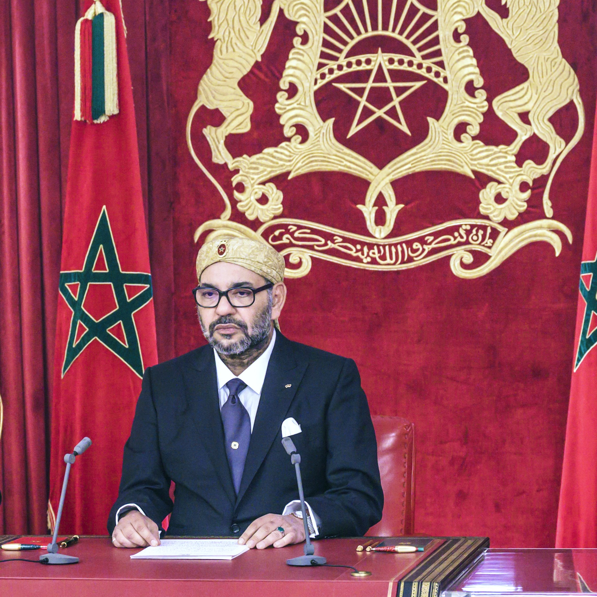 L'image montre un homme assis à un bureau, habillé élégamment en costume noir avec une cravate. Il porte un turban ou un couvre-chef traditionnel. En arrière-plan, on peut voir deux drapeaux marocains ornés d'une étoile à cinq branches. Le décor est riche, avec un fond rouge et une représentation du blason du Maroc. L'homme semble concentré et prêt à s'exprimer.