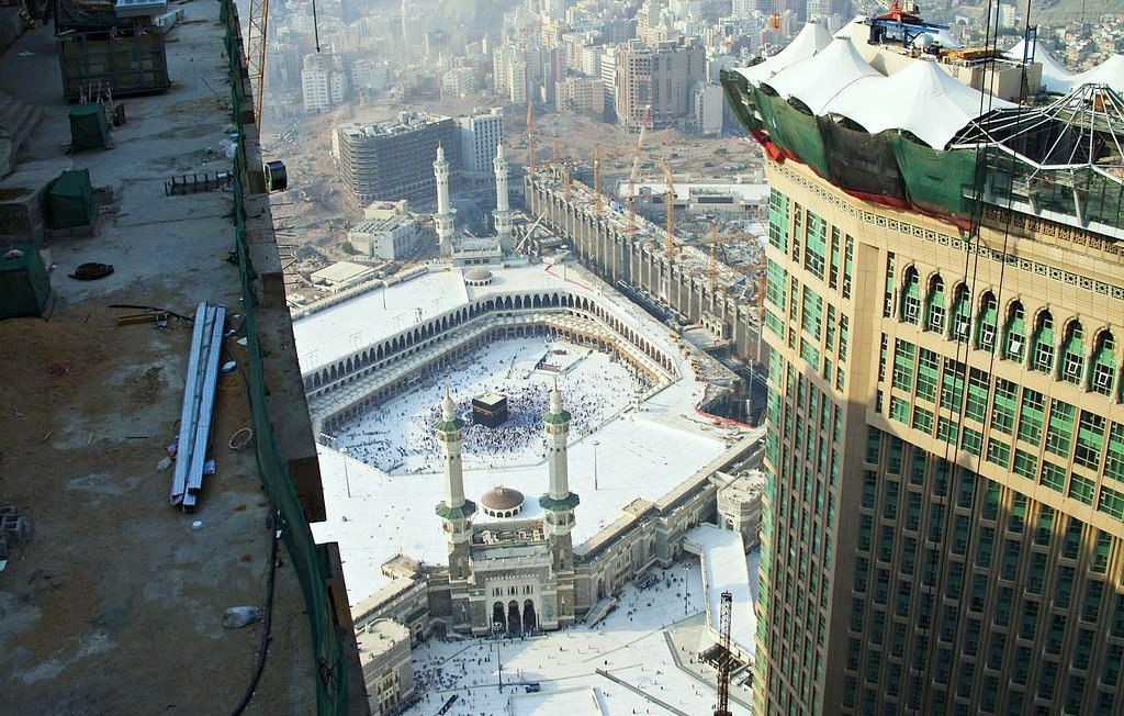 L'image montre une vue aérienne de la Kaaba à La Mecque, en Arabie Saoudite. On peut voir le complexe du Haram qui comprend la mosquée entourant la Kaaba. Il y a de nombreux pèlerins qui s'y trouvent, et l'environnement urbain avec des bâtiments modernes en arrière-plan. On aperçoit également des travaux de construction en cours sur le côté, suggérant une expansion des infrastructures autour du site sacré. Le ciel est légèrement brumeux, ce qui donne une ambiance particulière à la scène.