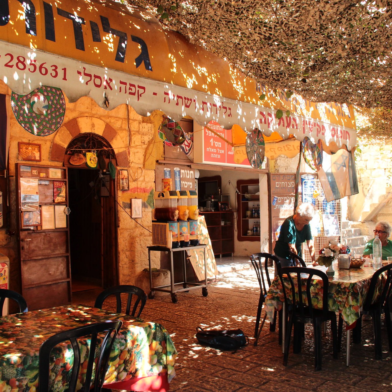 La imagen muestra un ambiente acogedor y colorido de un café o restaurante al aire libre. Hay mesas y sillas negras distribuidas sobre un suelo pavimentado, y la decoración incluye manteles con estampados florales. En el fondo, se pueden ver varias banderas, incluyendo la de Israel, colgadas en las paredes de piedra. El espacio está cubierto por una especie de toldo que filtra la luz, creando un ambiente agradable. Algunas personas están sentadas en las mesas, disfrutando del lugar.