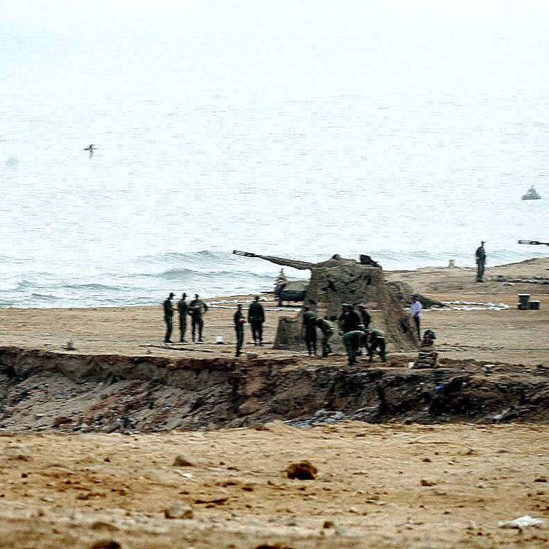 L'image montre une scène côtière où des militaires sont visibles sur une plage. On aperçoit des canons posés sur le sable, ainsi qu'un groupe de soldats qui semblent s'affairer autour. Le paysage est brumeux, avec une mer calme au fond et un ciel nuageux. L'atmosphère paraît sérieuse et militaire, avec une concentration sur les préparatifs ou les opérations en cours.